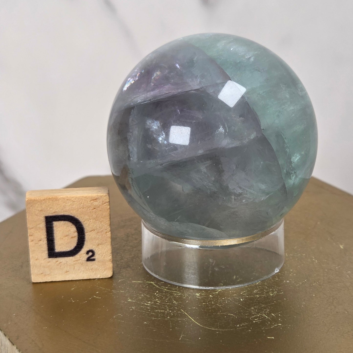 A polished round crystal sphere with green and purple tones sits on a clear acrylic stand next to a wooden Scrabble tile showing the letter D, set on a gold and wood pedestal.