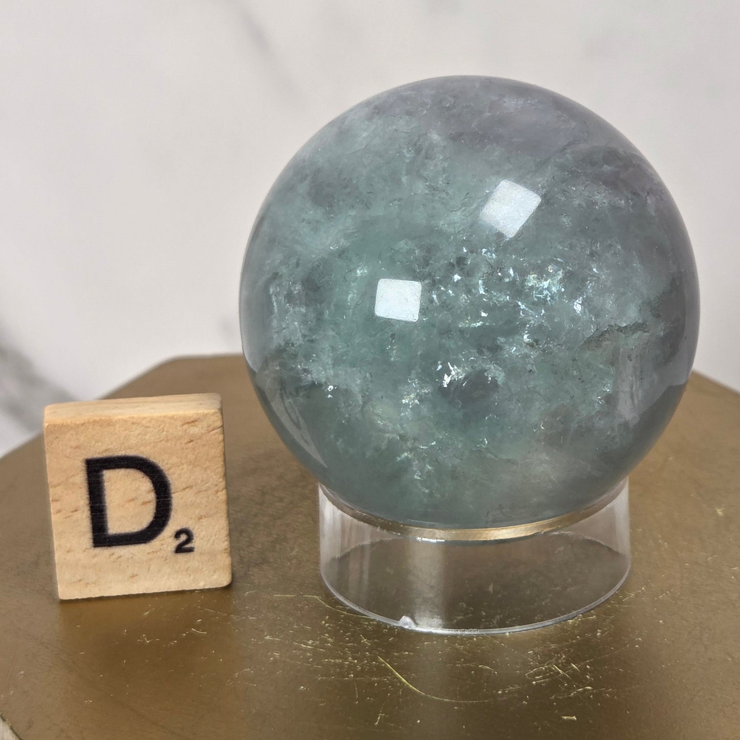 A polished round crystal sphere in a muted blue-green hue, displayed on a clear acrylic stand next to a wooden Scrabble tile on a gold hexagonal pedestal.