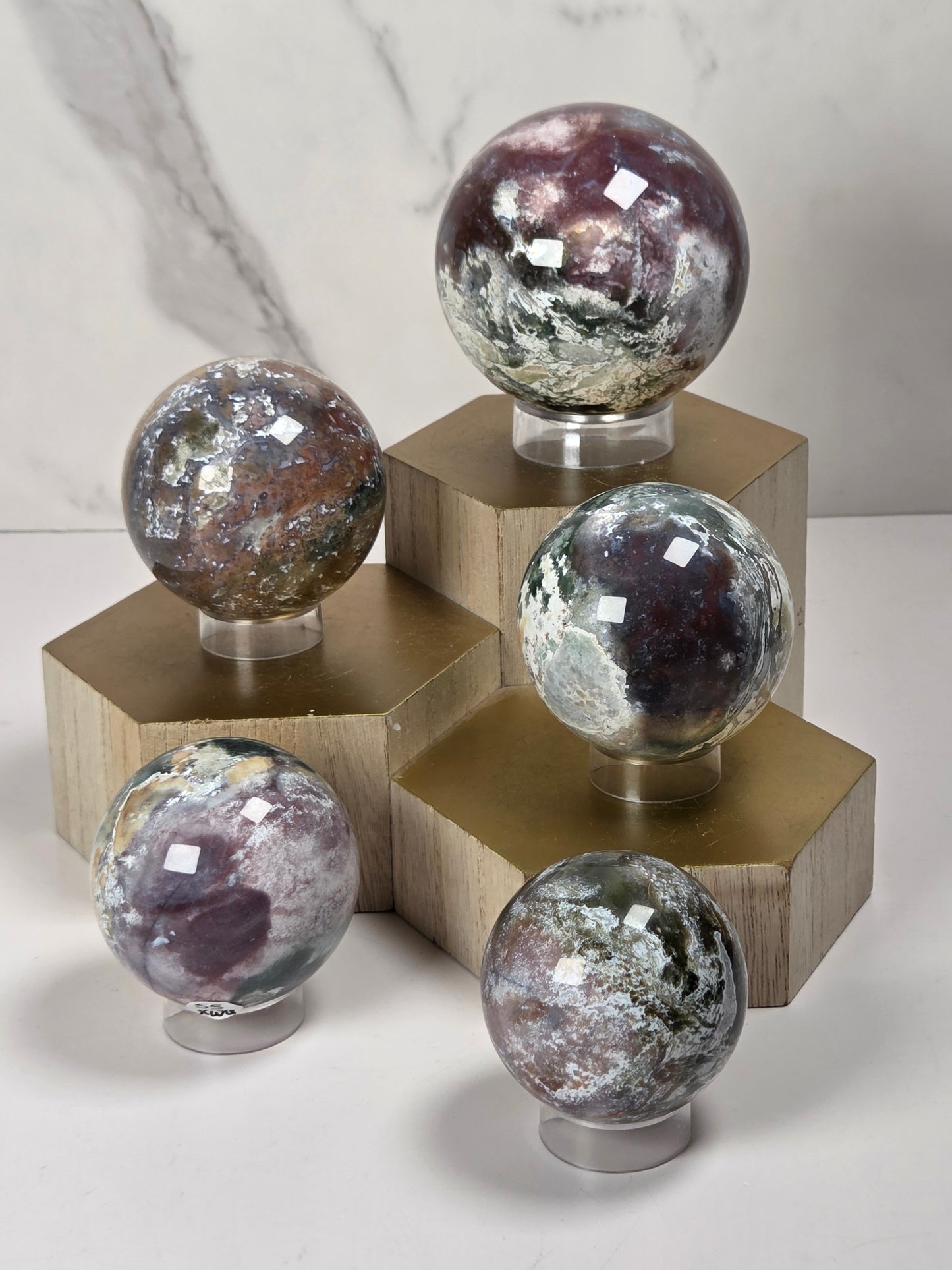 Five polished crystal spheres with natural patterns and metallic sheen, each mounted on a clear stand and placed on a gold-colored geometric display block against a light marble background.