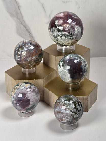 Five polished crystal spheres with natural patterns and metallic sheen, each mounted on a clear stand and placed on a gold-colored geometric display block against a light marble background.