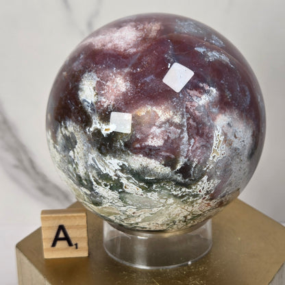 A polished natural crystal sphere with marbled purple, white, and green patterns, displayed on a clear stand atop a gold hexagonal pedestal. A small wooden block with the letter A is placed beside it against a light marble-patterned background.