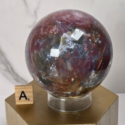 A polished round stone sphere with marbled burgundy, brown, gray, and white patterns, displayed on a clear stand atop a gold-colored wooden pedestal. A small wooden letter tile with the letter 'A' sits next to the base.