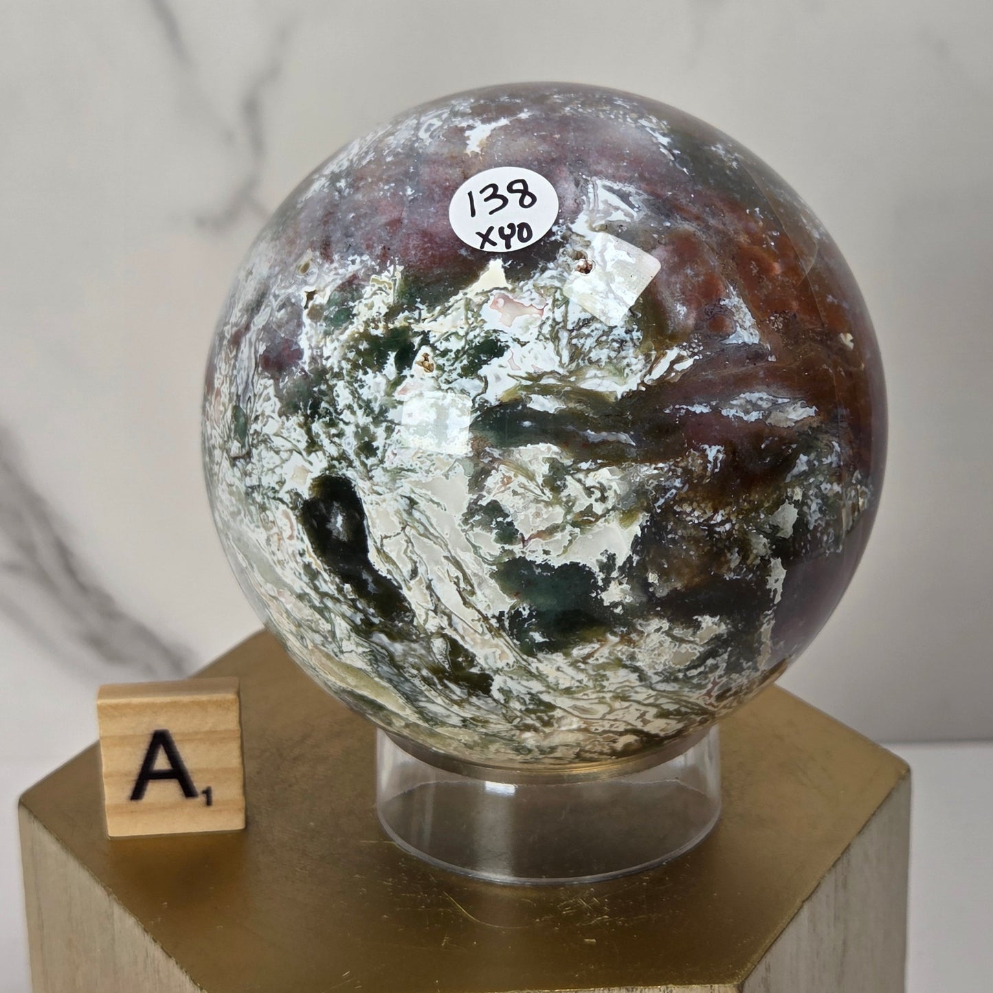 A polished round stone sphere with marbled brown, white, and dark green patterns, displayed on a clear stand atop a gold-colored block pedestal. A small wooden letter tile showing 'A1' sits beside it against a light marble-patterned background.