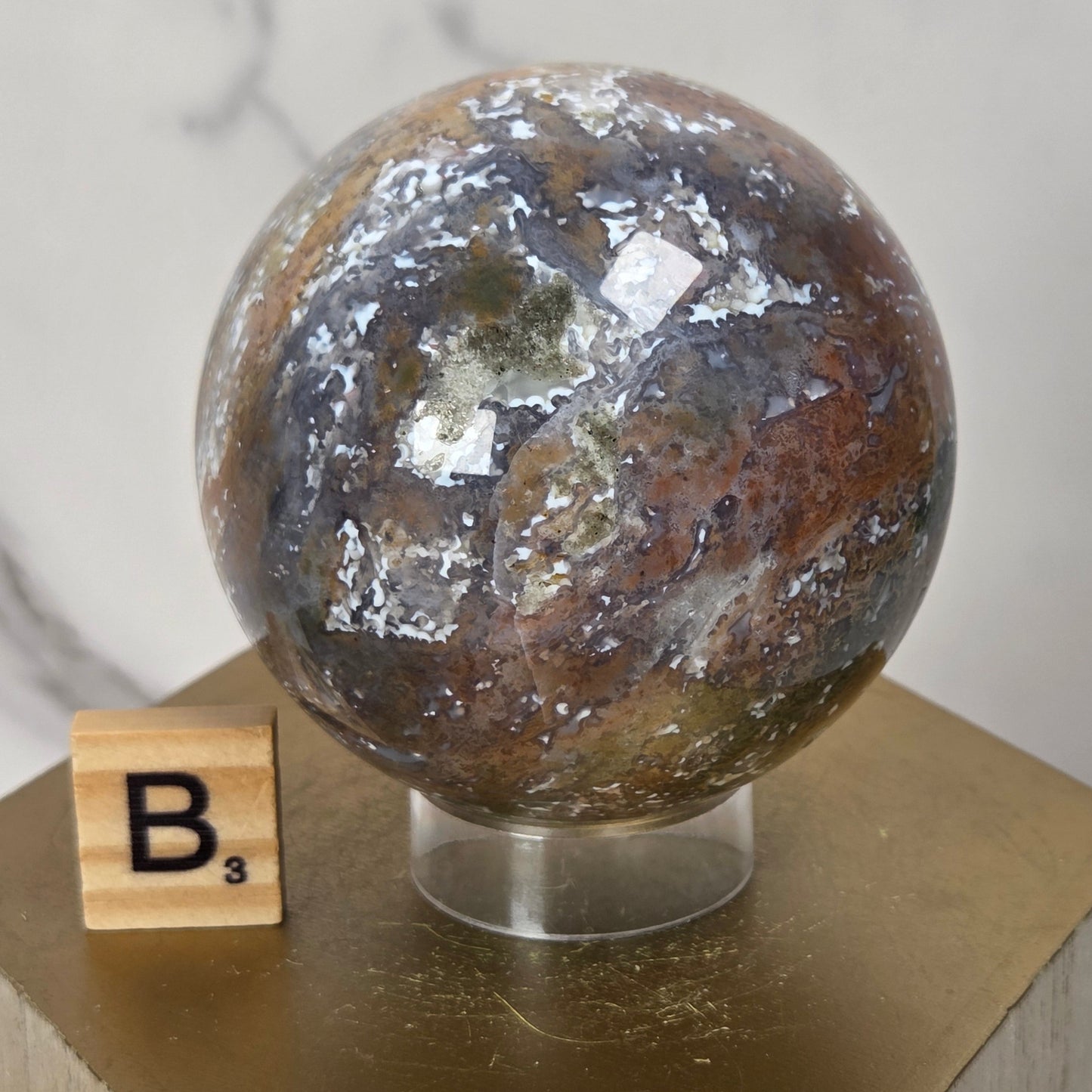 A polished round stone sphere with marbled brown, white, and dark green patterns, displayed on a clear stand atop a gold-colored block pedestal. A small wooden letter tile showing 'B1' sits beside it against a light marble-patterned background.