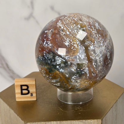A polished decorative crystal sphere with swirling brown, white, gray, and dark green patterns, displayed on a clear stand atop a gold-toned pedestal next to a wooden Scrabble tile.