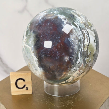 A polished geode sphere with a dark brown center and swirling green, gray, and white outer layers, displayed on a clear stand atop a gold hexagonal pedestal next to a Scrabble tile.