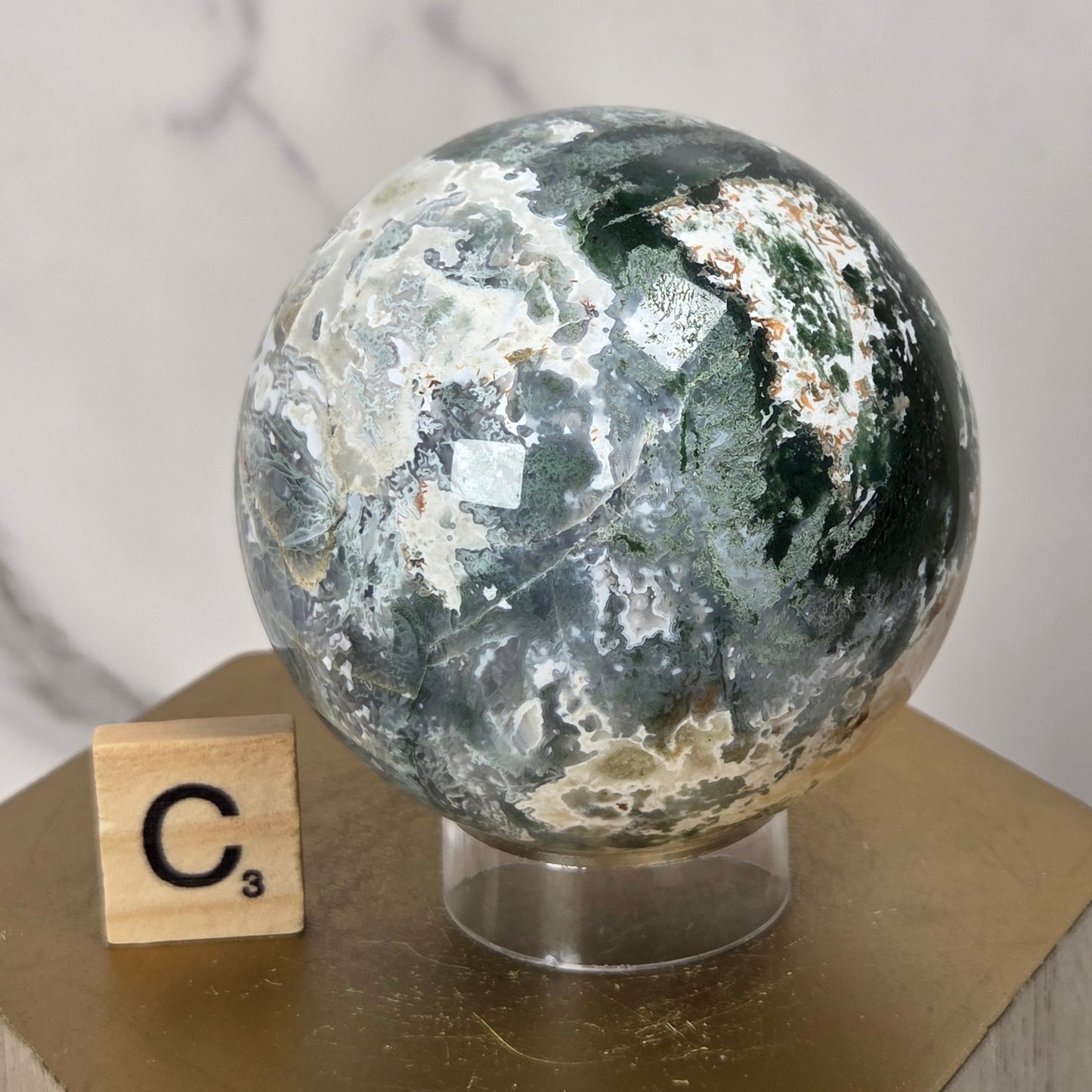 A polished natural crystal sphere with green, white, and gray marbling, displayed on a clear stand atop a gold hexagonal pedestal next to a Scrabble tile.