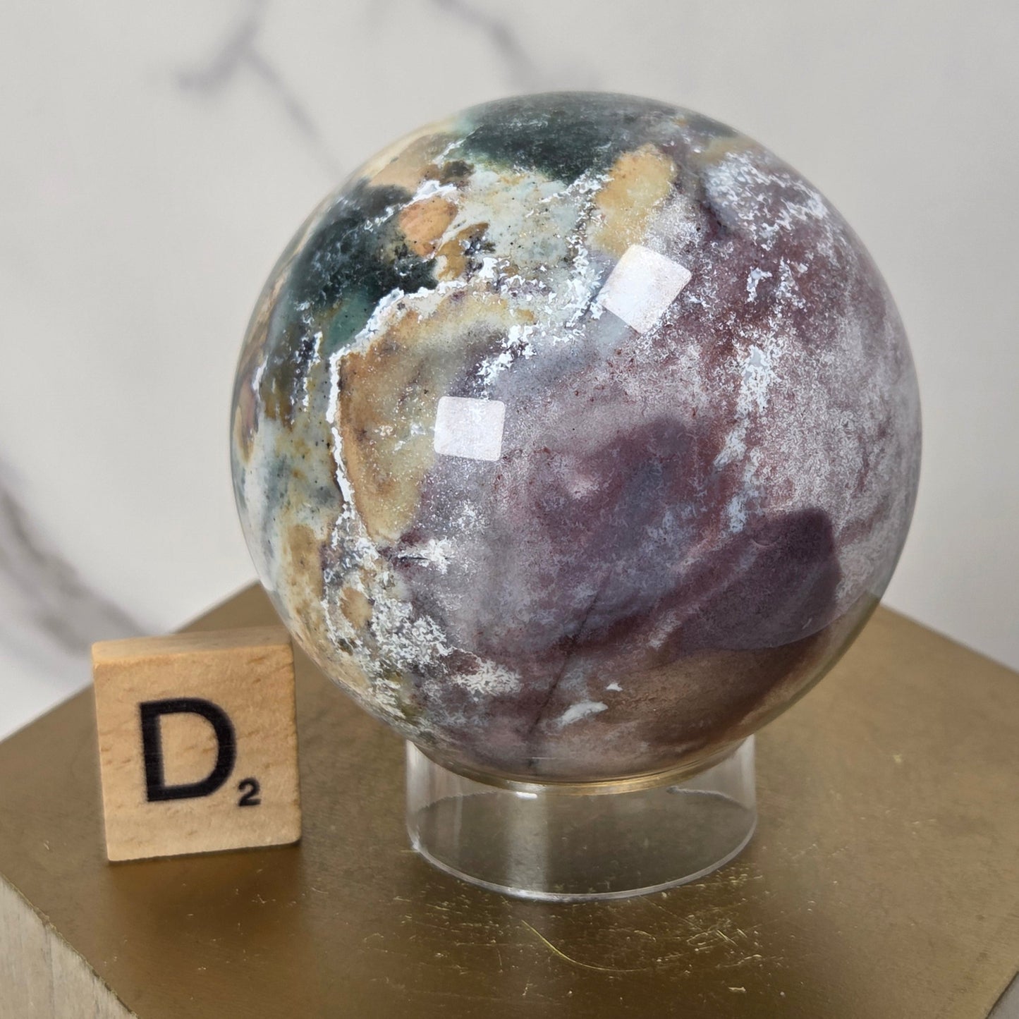 A polished natural stone sphere with marbled green, brown, beige, and white patterns, displayed on a clear stand atop a gold hexagonal pedestal next to a wooden Scrabble tile.