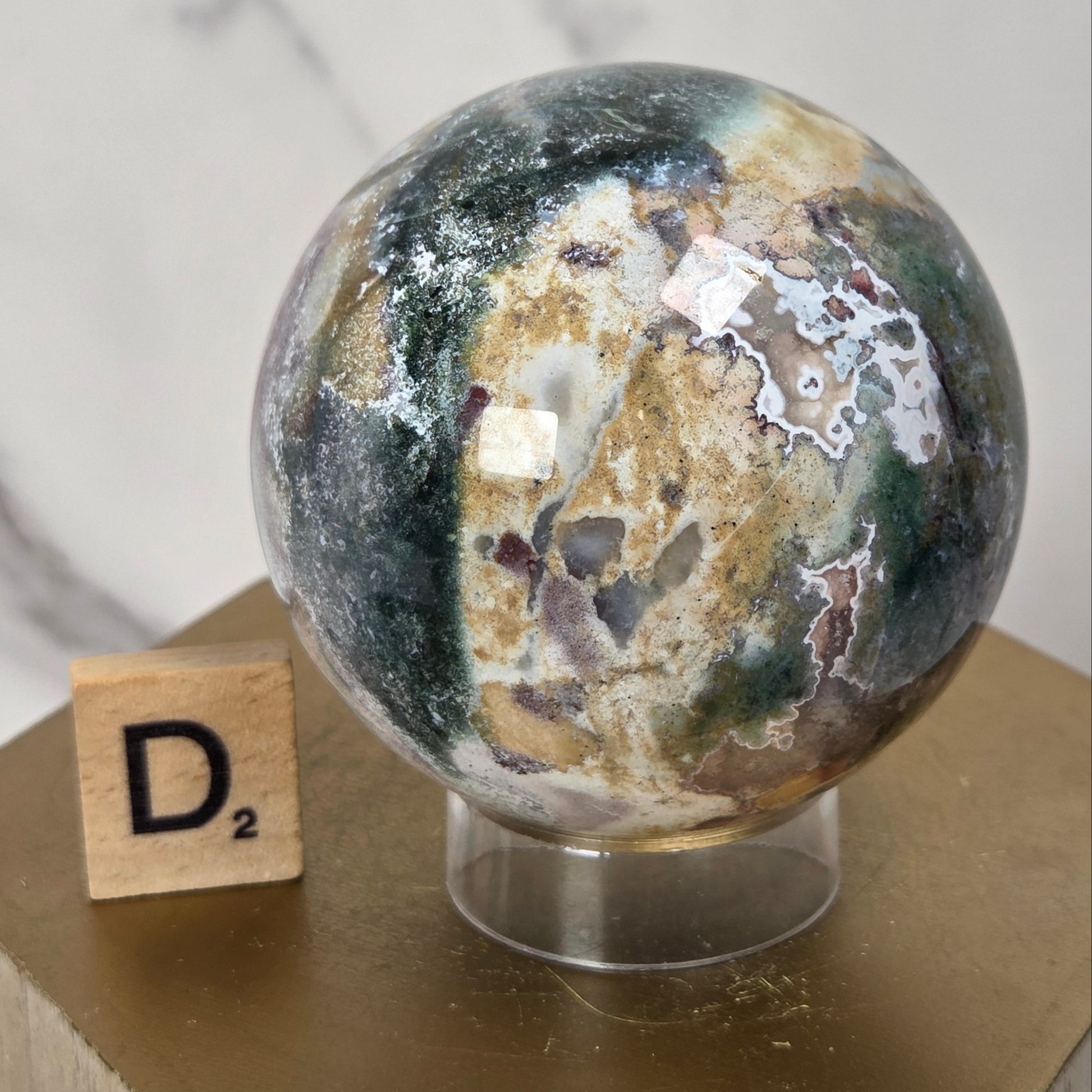 A polished natural stone sphere with marbled green, brown, beige, and white patterns, displayed on a clear stand atop a gold hexagonal pedestal next to a wooden Scrabble tile.