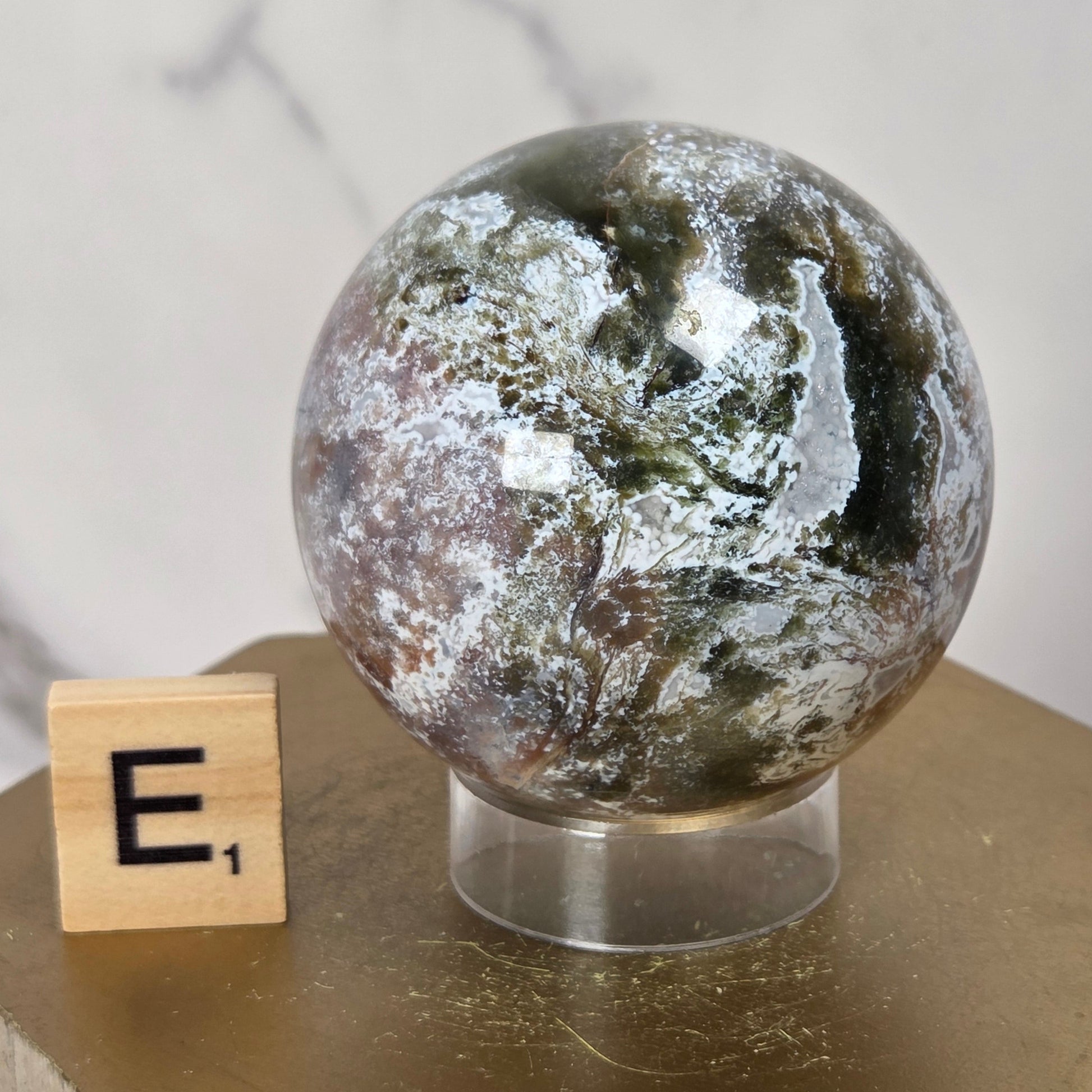 A polished natural crystal sphere with earthy green, brown, gray, and white marbling, displayed on a clear acrylic stand atop a gold hexagonal pedestal. A small wooden letter tile showing 'E1' sits beside it against a light marble-patterned background.