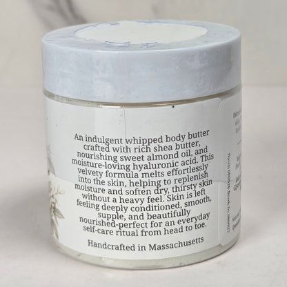 A white jar of whipped body butter with a white lid and black text on the label, set against a soft, light gray background.