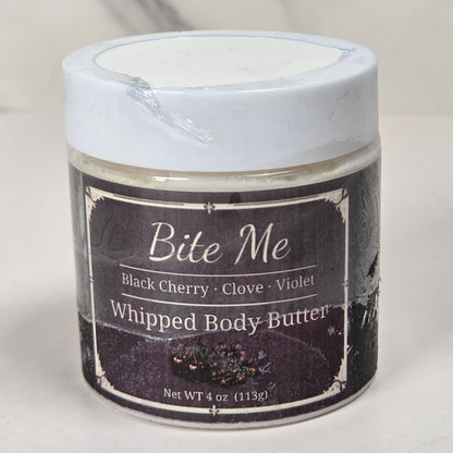 A jar of Bite Me Whipped Body Butter with black cherry, clove, and violet ingredients, featuring a white lid and a dark label, set against a light marble background.