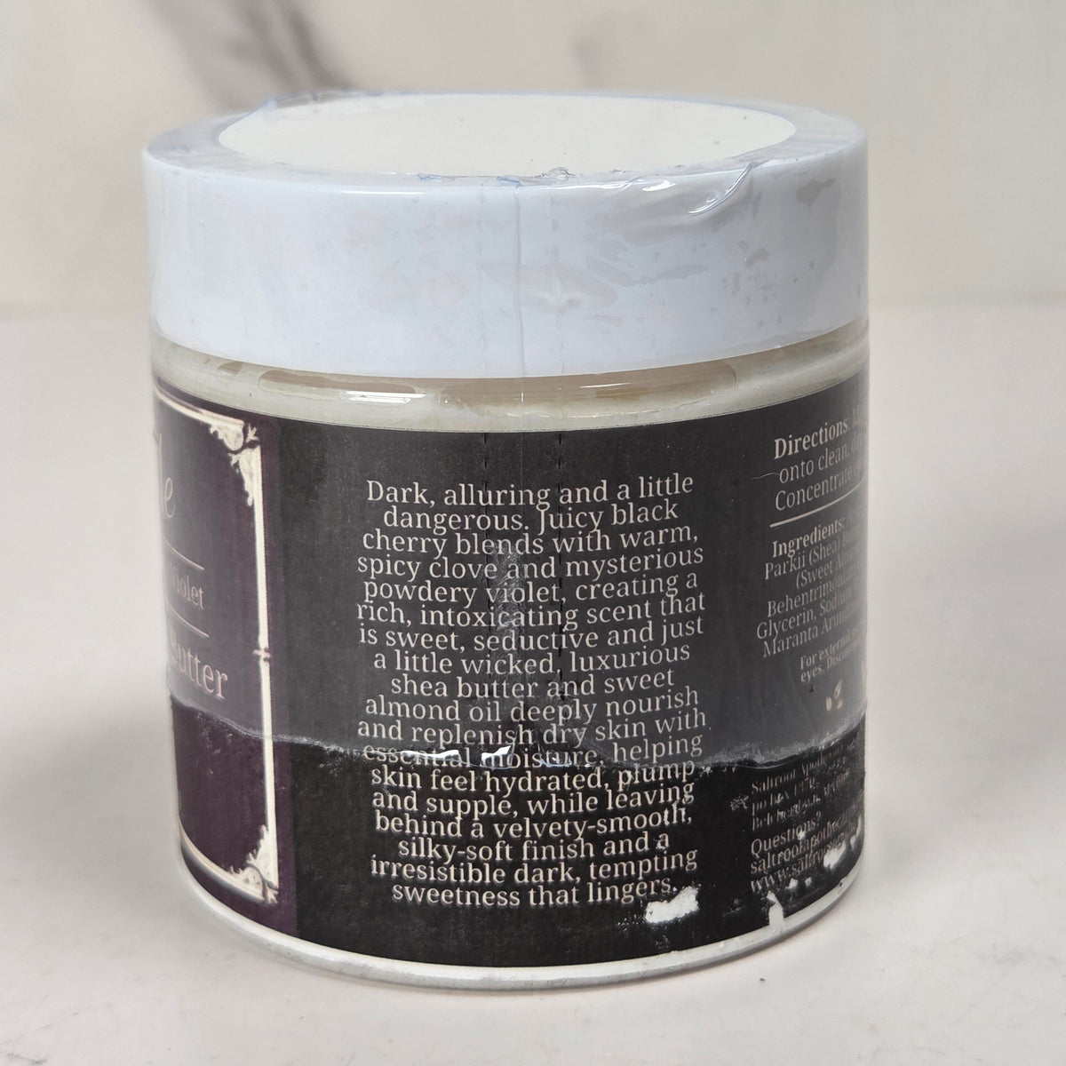 A jar of whipped body butter with a white lid and a printed product description label, photographed against a light neutral background.