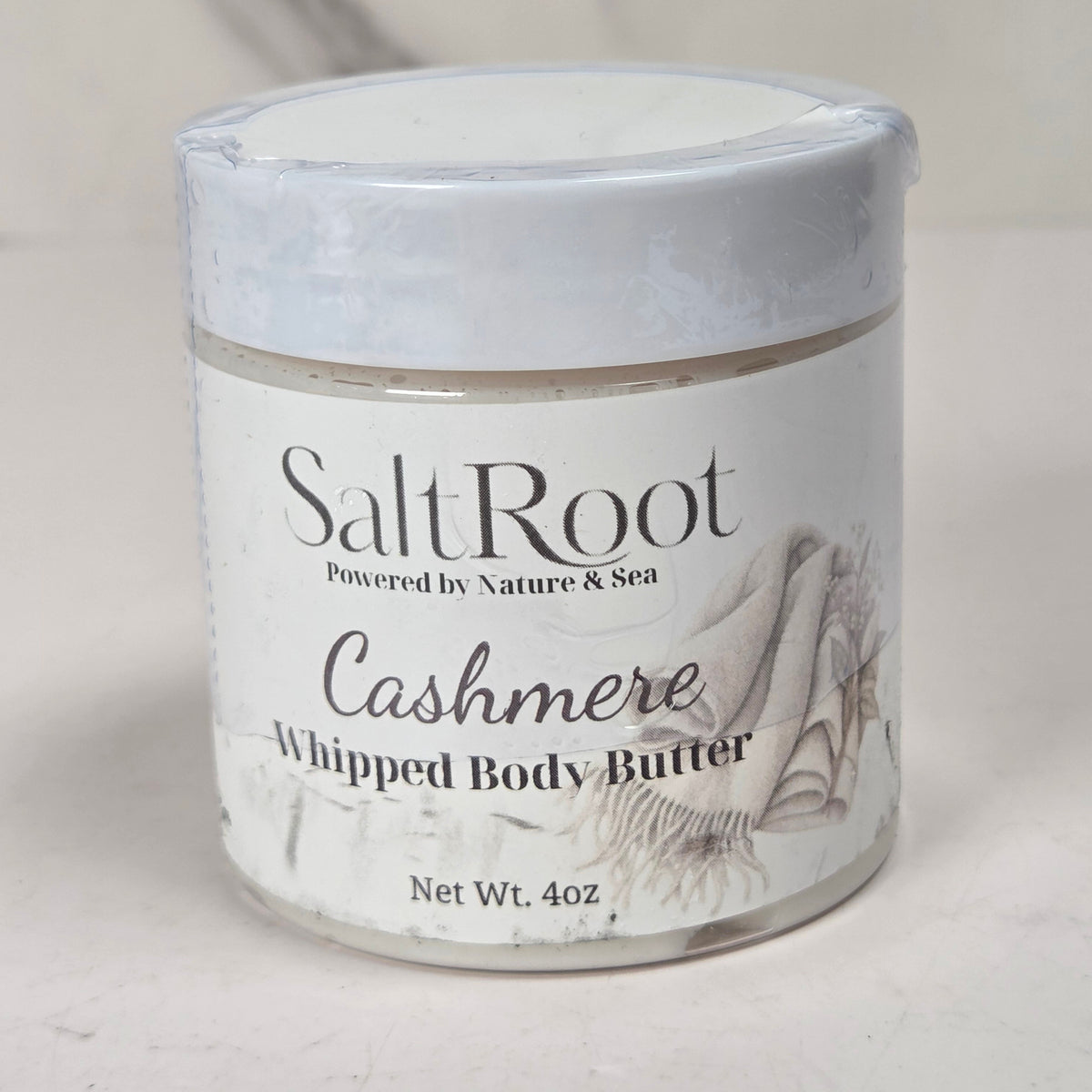 A 4 oz jar of SaltRoot Cashmere Whipped Body Butter, featuring a white label with black text and a seashell illustration, set against a neutral light gray background.