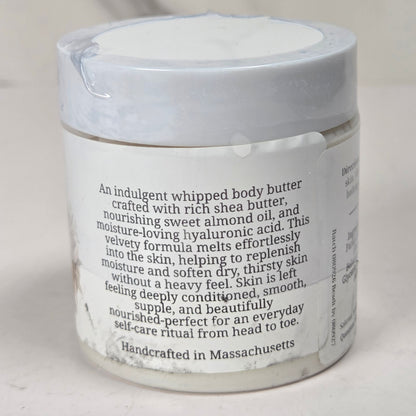 A white jar of whipped body butter with a white lid and a printed product description label, photographed against a light neutral background.