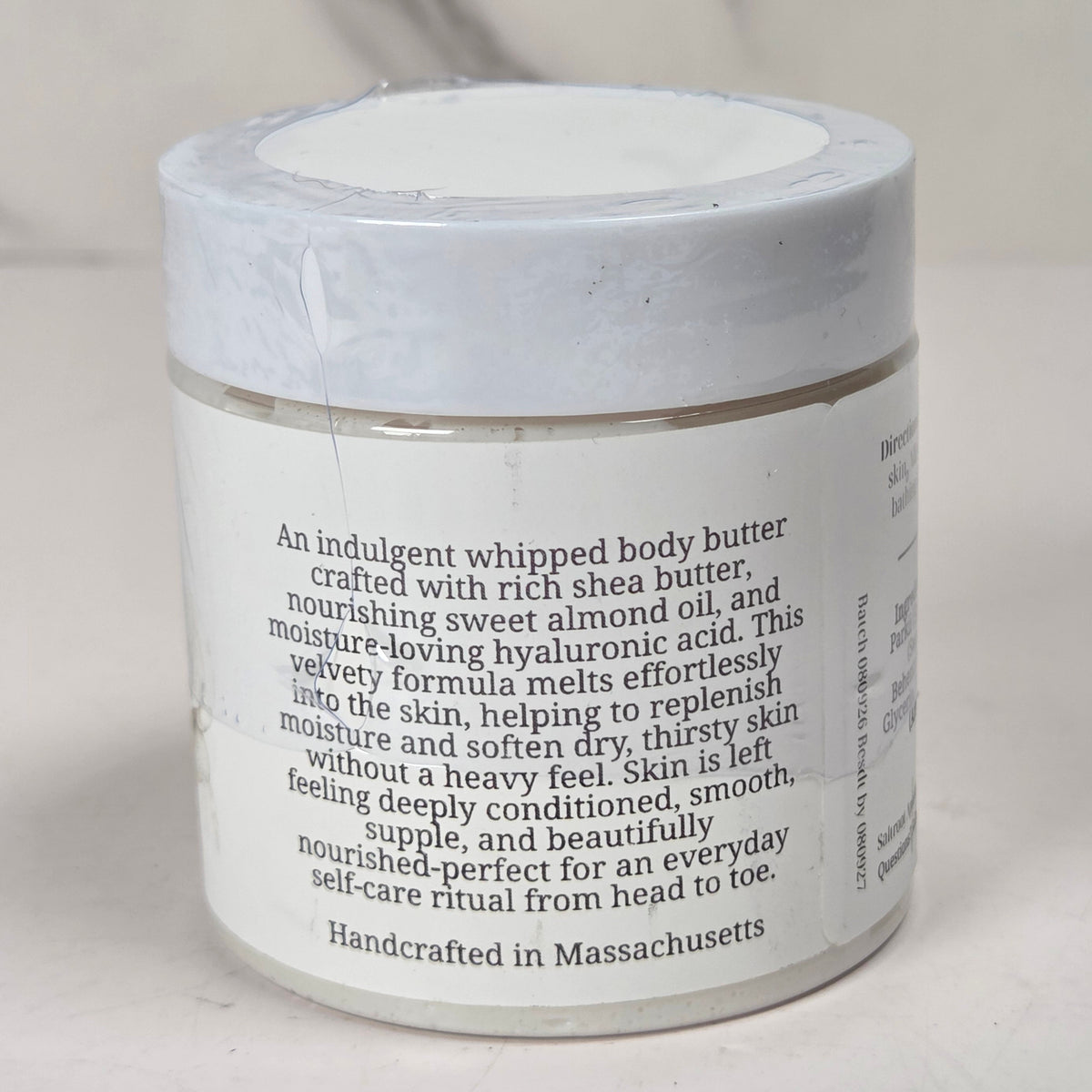 A white jar of whipped body butter with a white lid and a white label featuring black descriptive text, set against a neutral light gray background.