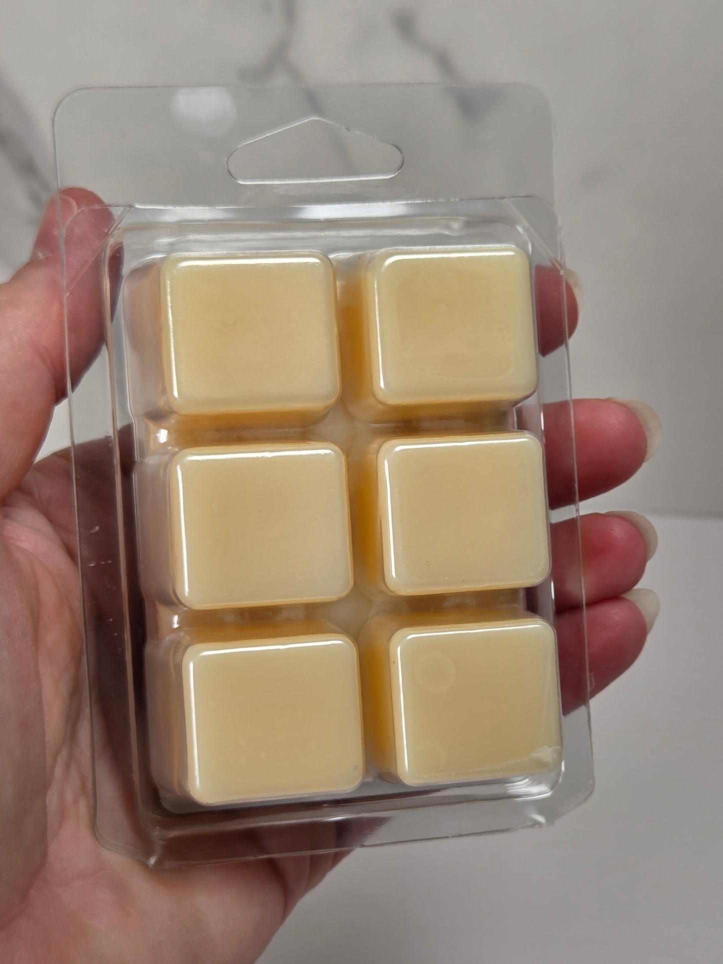 A hand holding a clear plastic blister pack containing six pale yellow cube-shaped wax melts or soap cubes against a light gray marble-patterned background.