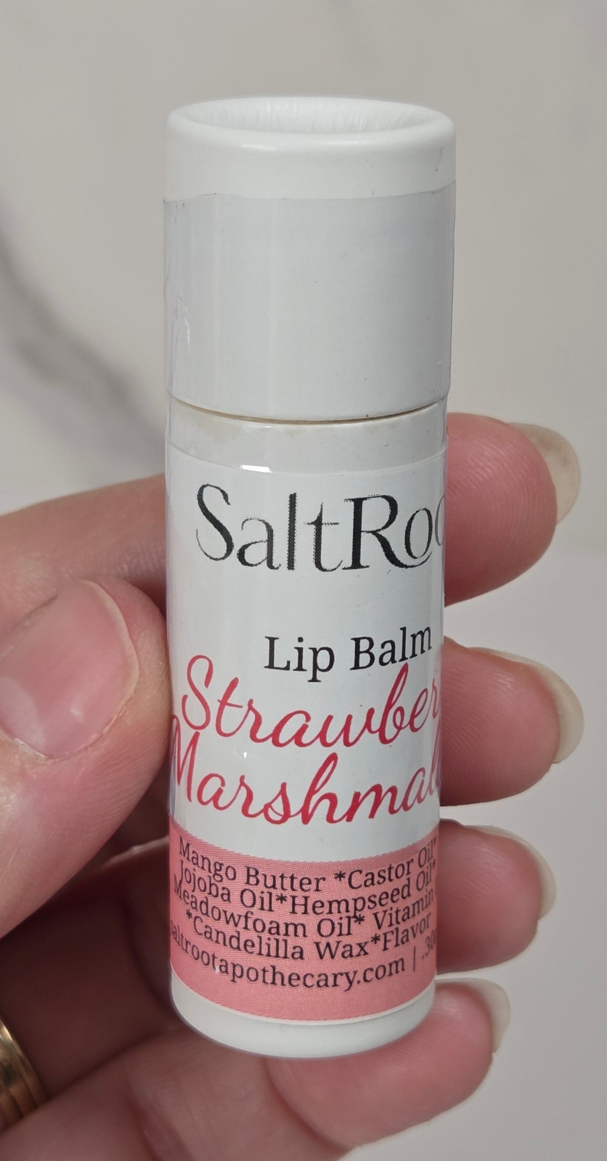 A hand holding a small white tube of Salt Ro Strawberry Marshmallow lip balm against a soft light gray marble background.