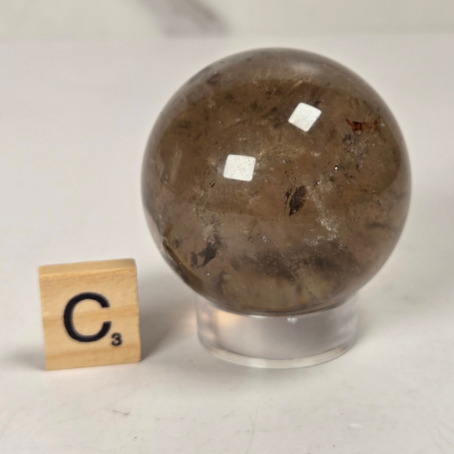 A polished brown quartz crystal sphere on a clear acrylic stand, shown next to a wooden Scrabble tile spelling 'C3' on a light gray surface.