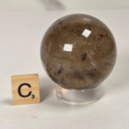 A polished brown quartz crystal sphere on a clear acrylic stand, shown next to a wooden Scrabble tile spelling 'C3' on a light gray surface.