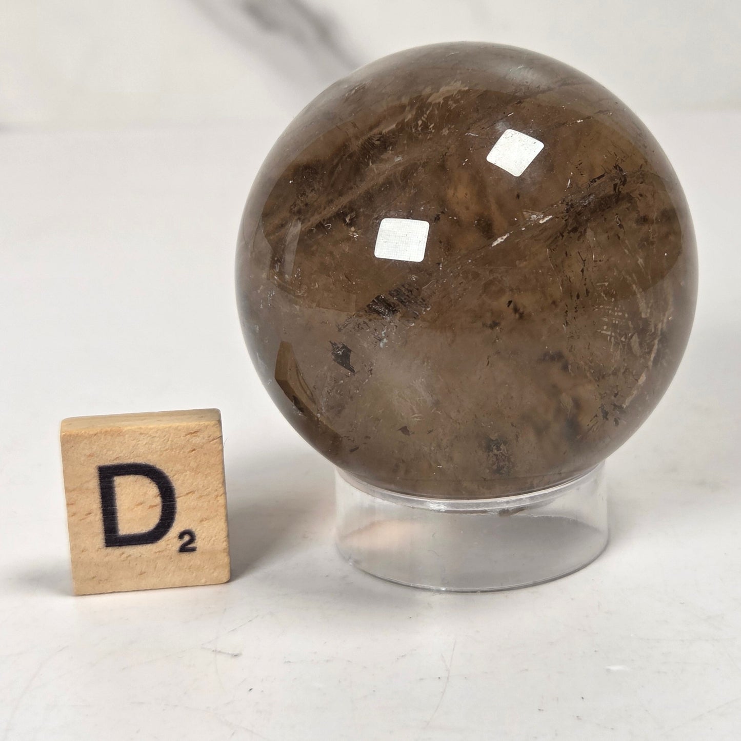 A polished brown smoky quartz crystal sphere on a clear acrylic stand, shown next to a wooden Scrabble tile spelling D2 on a light marble surface.