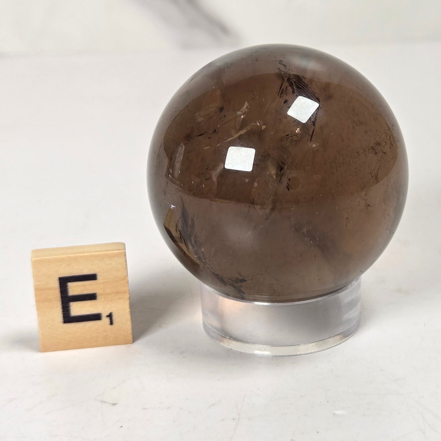 A polished brown smoky quartz crystal sphere displayed on a clear acrylic stand, with a small wooden Scrabble tile showing the letter 'E' placed to its left, set against a light gray background.