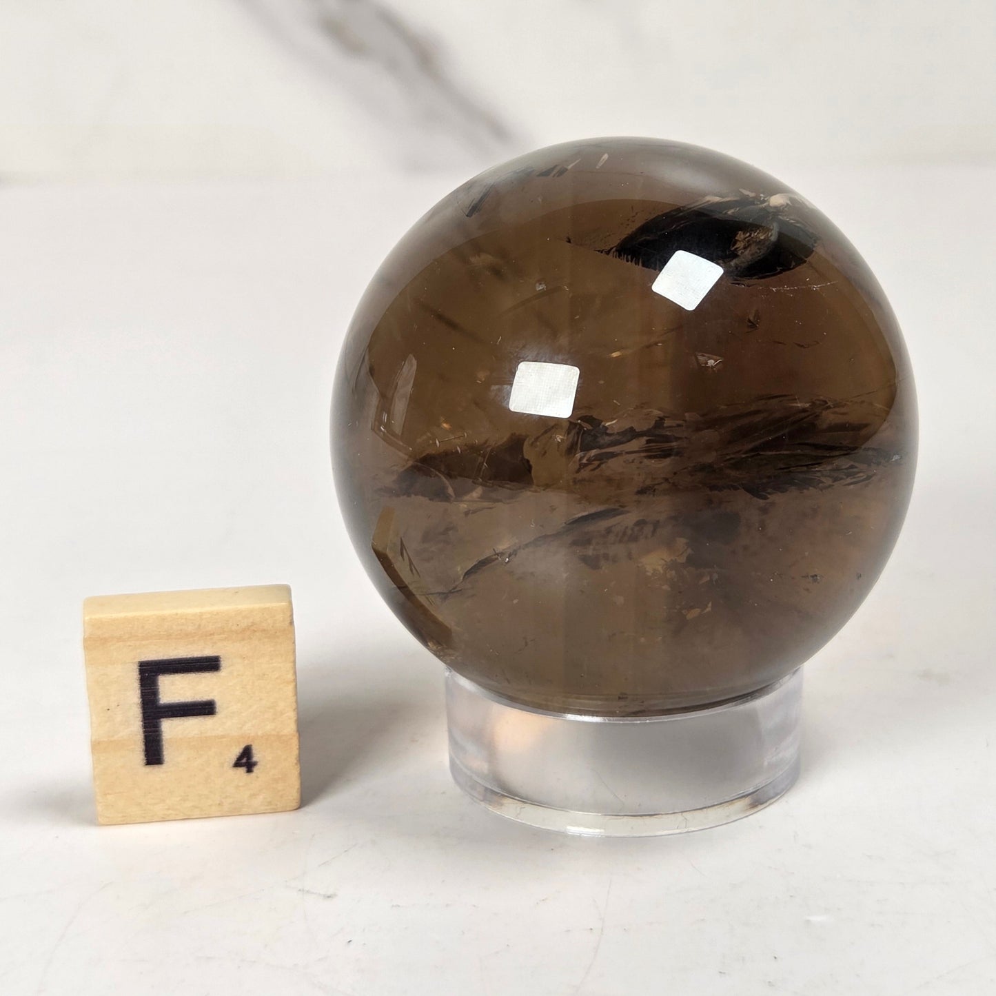 A polished brown smoky quartz crystal sphere on a clear acrylic stand, shown next to a wooden Scrabble tile with the letter F on a light marble surface.