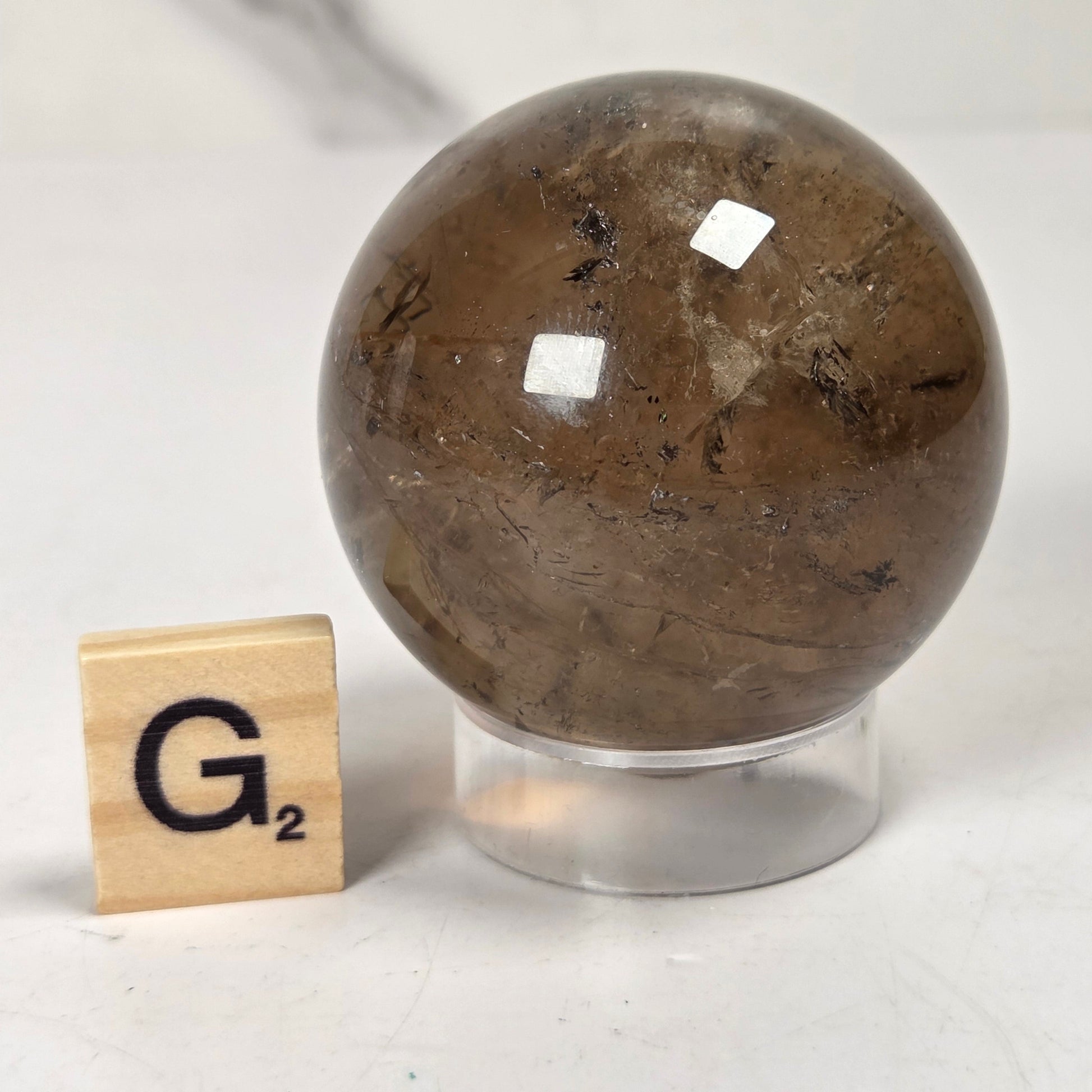 A polished brown quartz crystal sphere with visible inclusions, displayed on a clear acrylic stand next to a wooden Scrabble tile showing the letter G.