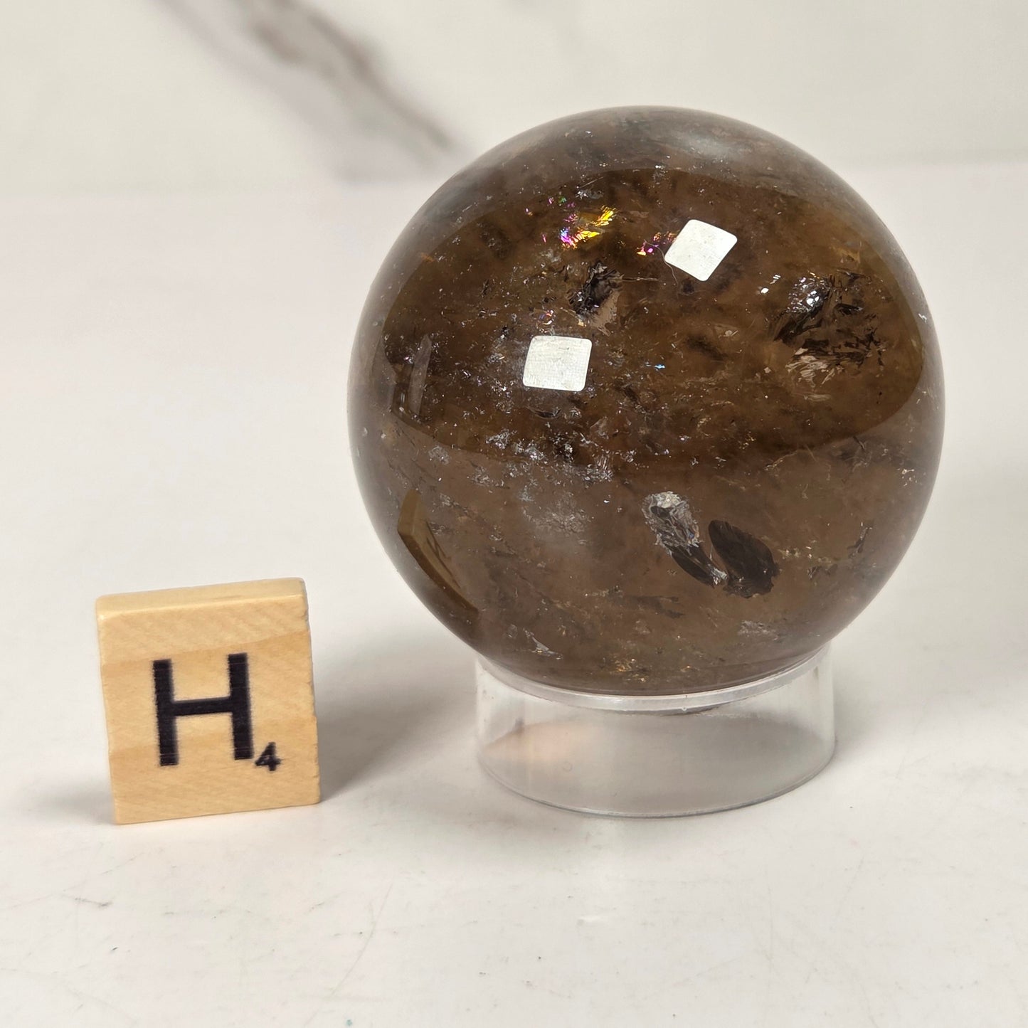 A polished brown crystal sphere with inclusions, displayed on a clear acrylic stand next to a small wooden Scrabble tile showing the letter H, on a light marble surface.