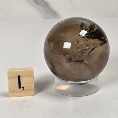 A smoky brown crystal sphere with natural inclusions, displayed on a clear acrylic stand next to a small wooden block with the number 1, on a light gray marble surface.