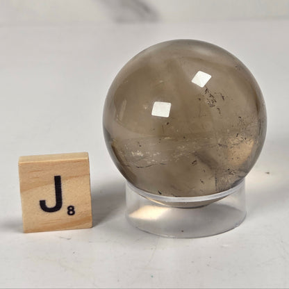 A smooth, polished smoky quartz crystal sphere on a clear acrylic stand, shown next to a wooden Scrabble tile spelling 'J8' on a light gray marble surface.