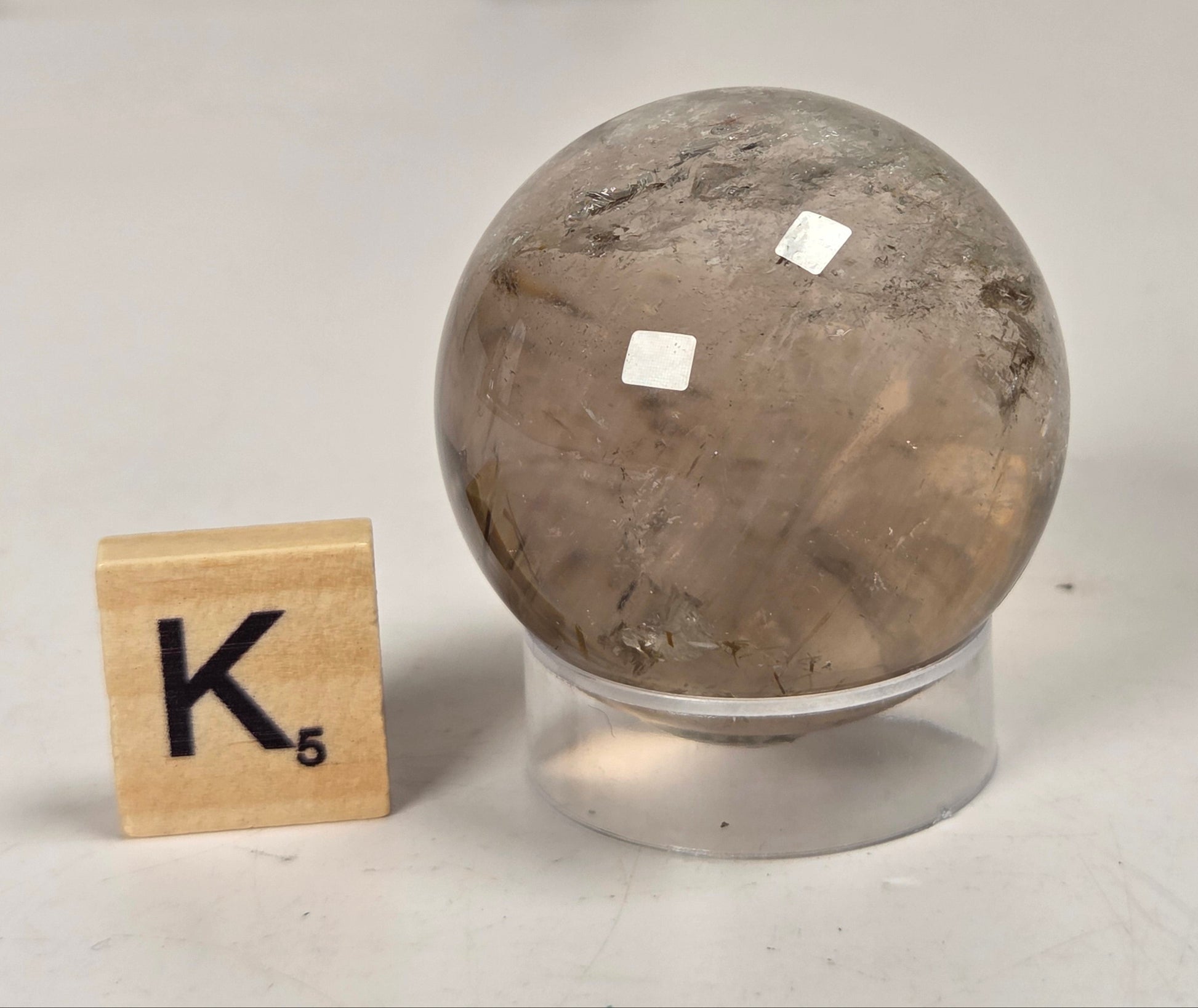 A polished crystal sphere with natural brown and gray marbling, displayed on a clear acrylic stand next to a wooden Scrabble tile showing the letter K.