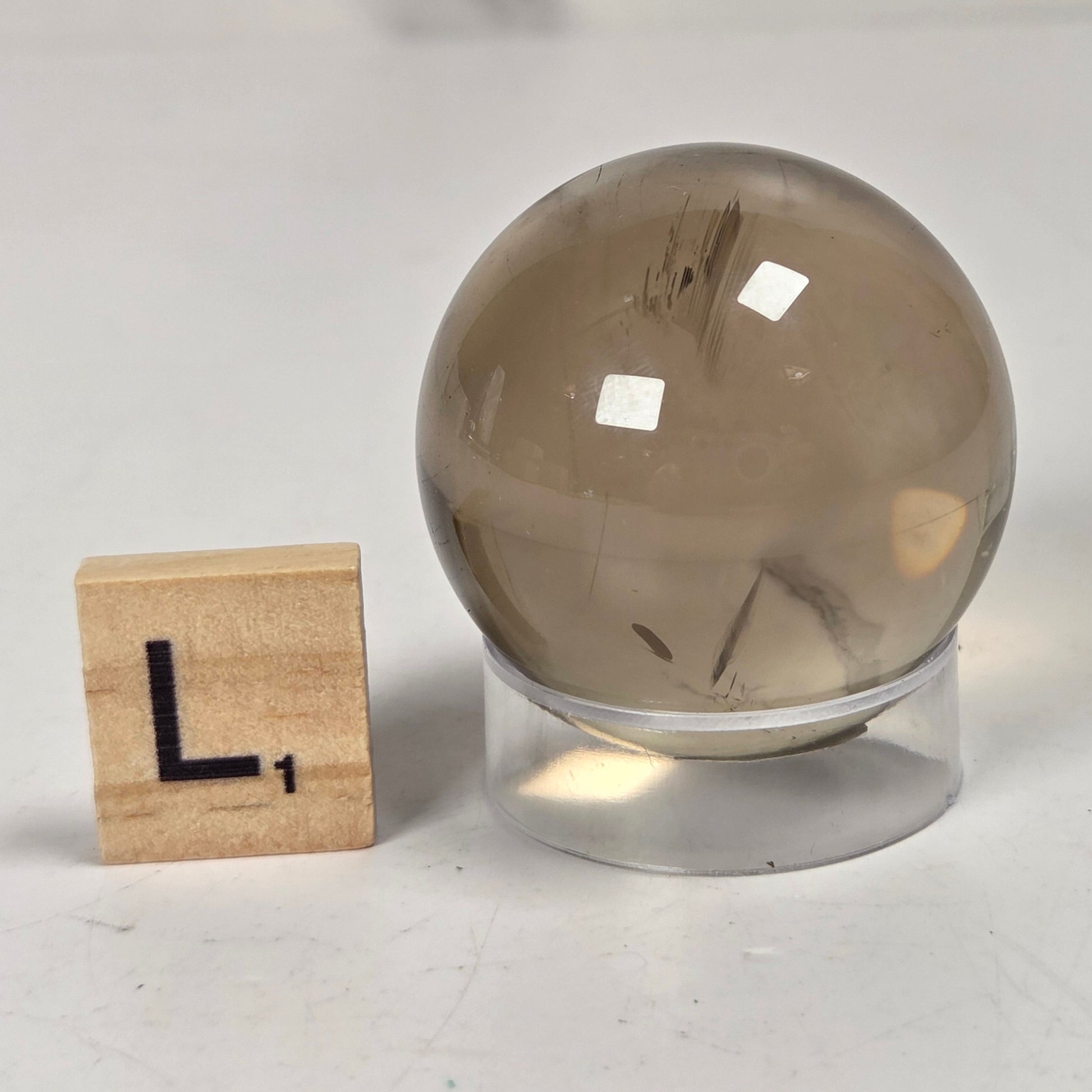 A smoky quartz crystal ball with visible inclusions, displayed on a clear acrylic stand next to a small wooden Scrabble tile showing the letter L.