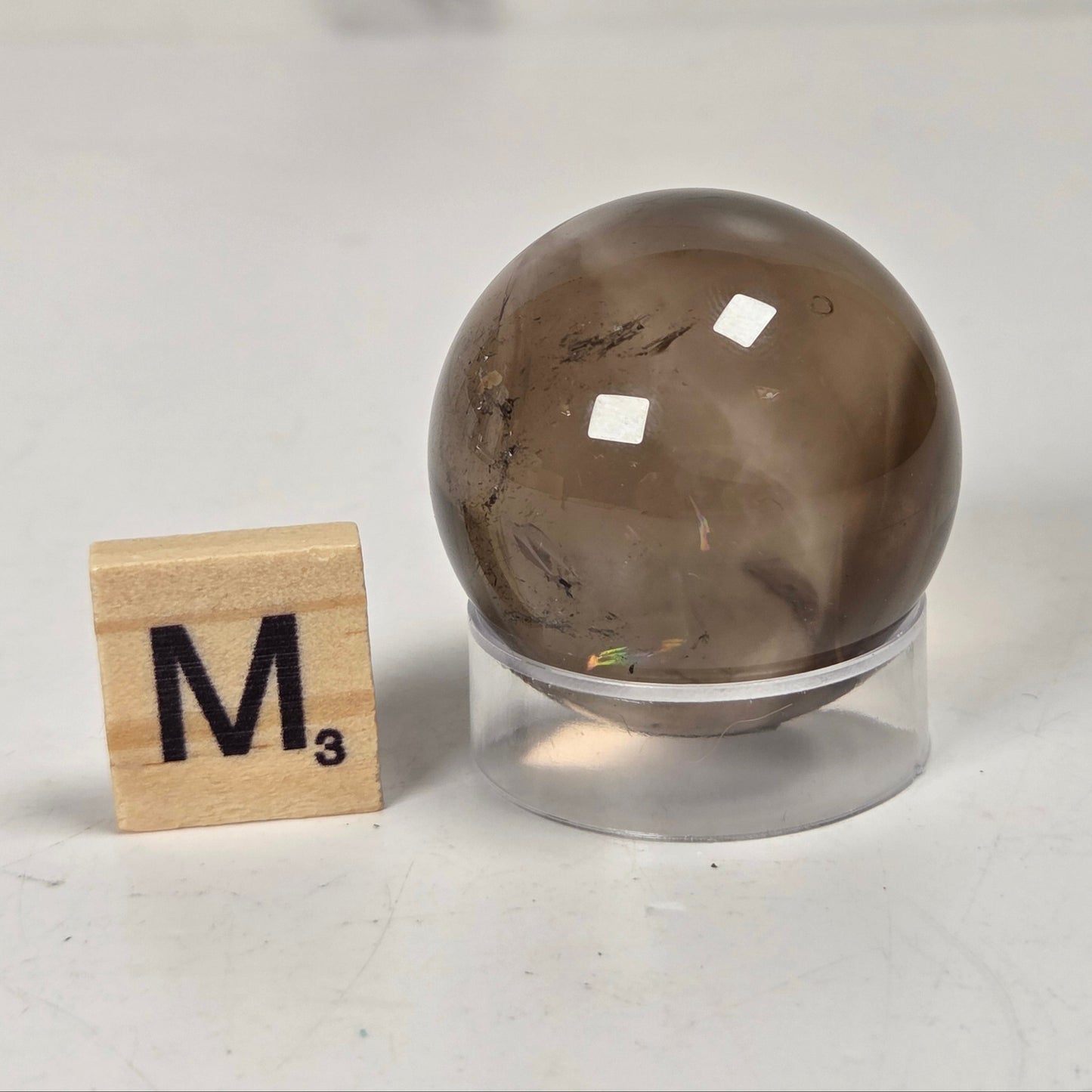 A polished brown smoky quartz crystal sphere on a clear acrylic stand, shown next to a wooden Scrabble tile with the letter M and subscript 3 on a light gray surface.