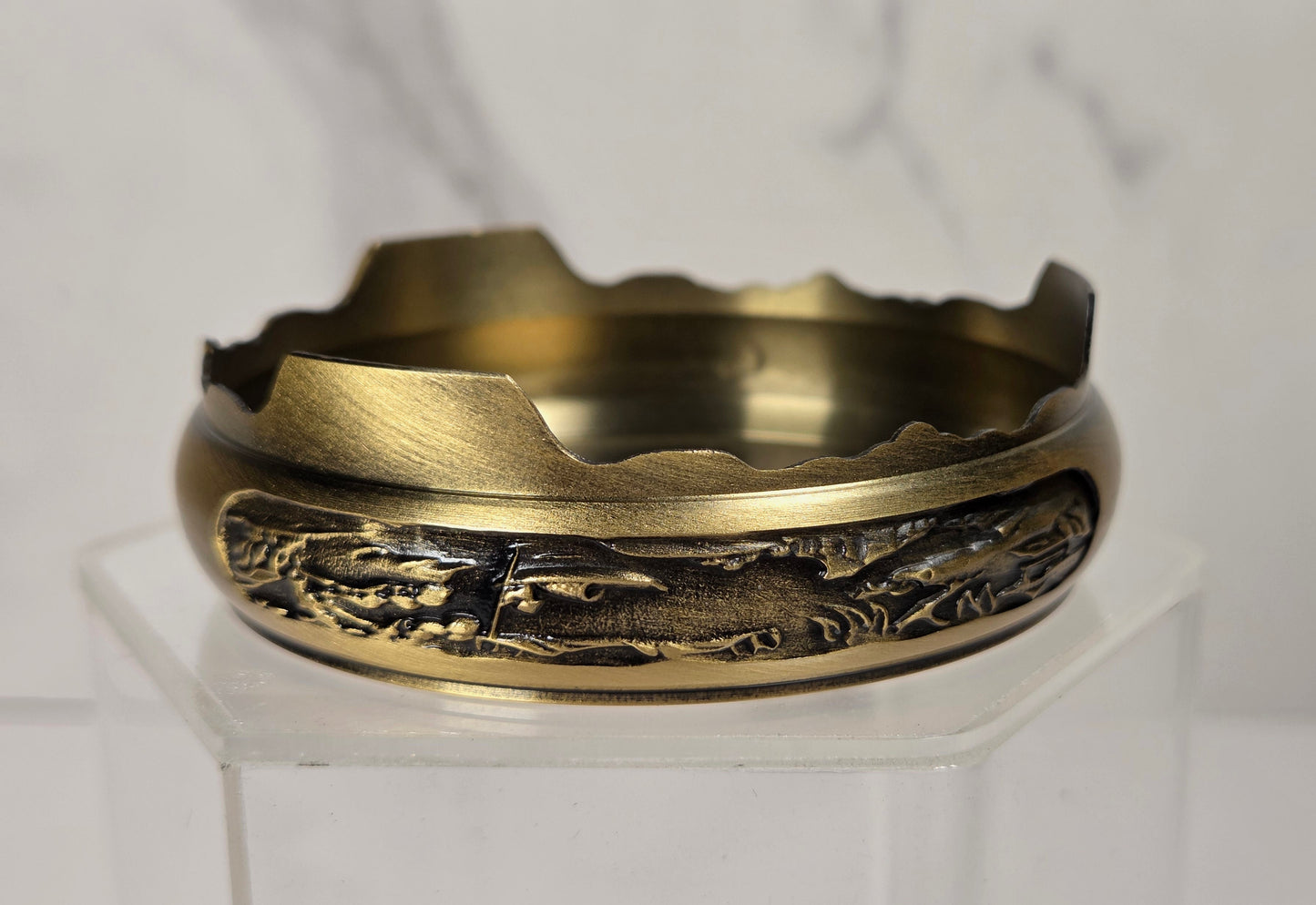 A vintage-style brass cigarette ashtray with a detailed landscape relief design on the front, displayed on a clear acrylic stand against a light gray marble background.