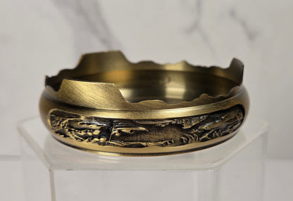 A vintage-style brass cigarette ashtray with a detailed landscape relief design on the front, displayed on a clear acrylic stand against a light gray marble background.