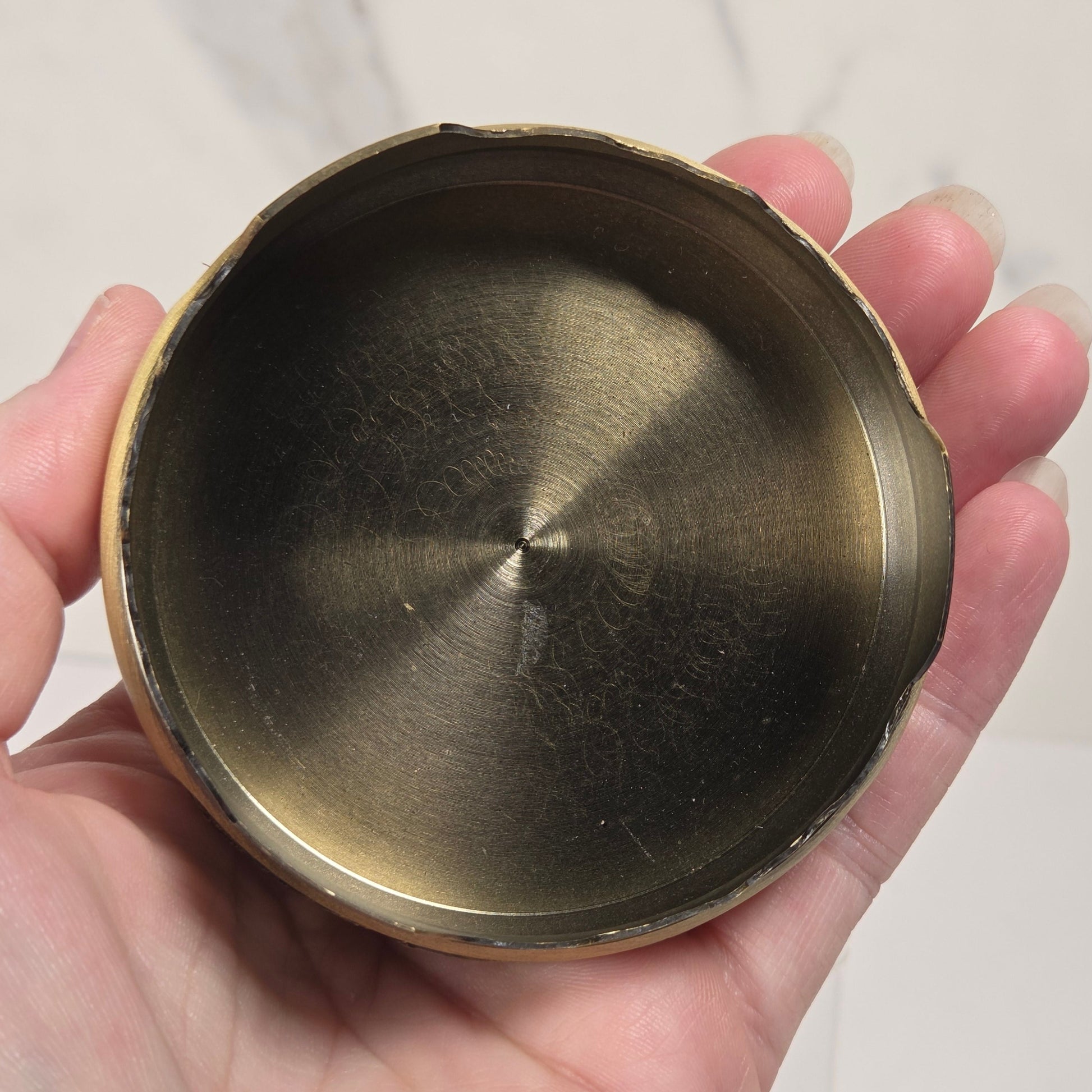 A hand holding a round, shallow brass container or dish with a brushed metallic interior and a slightly rough, rustic rim, set against a light marble-patterned background.