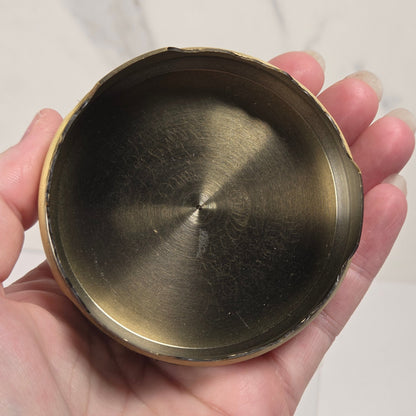 A hand holding a round, shallow brass container or dish with a brushed metallic interior and a slightly rough, rustic rim, set against a light marble-patterned background.