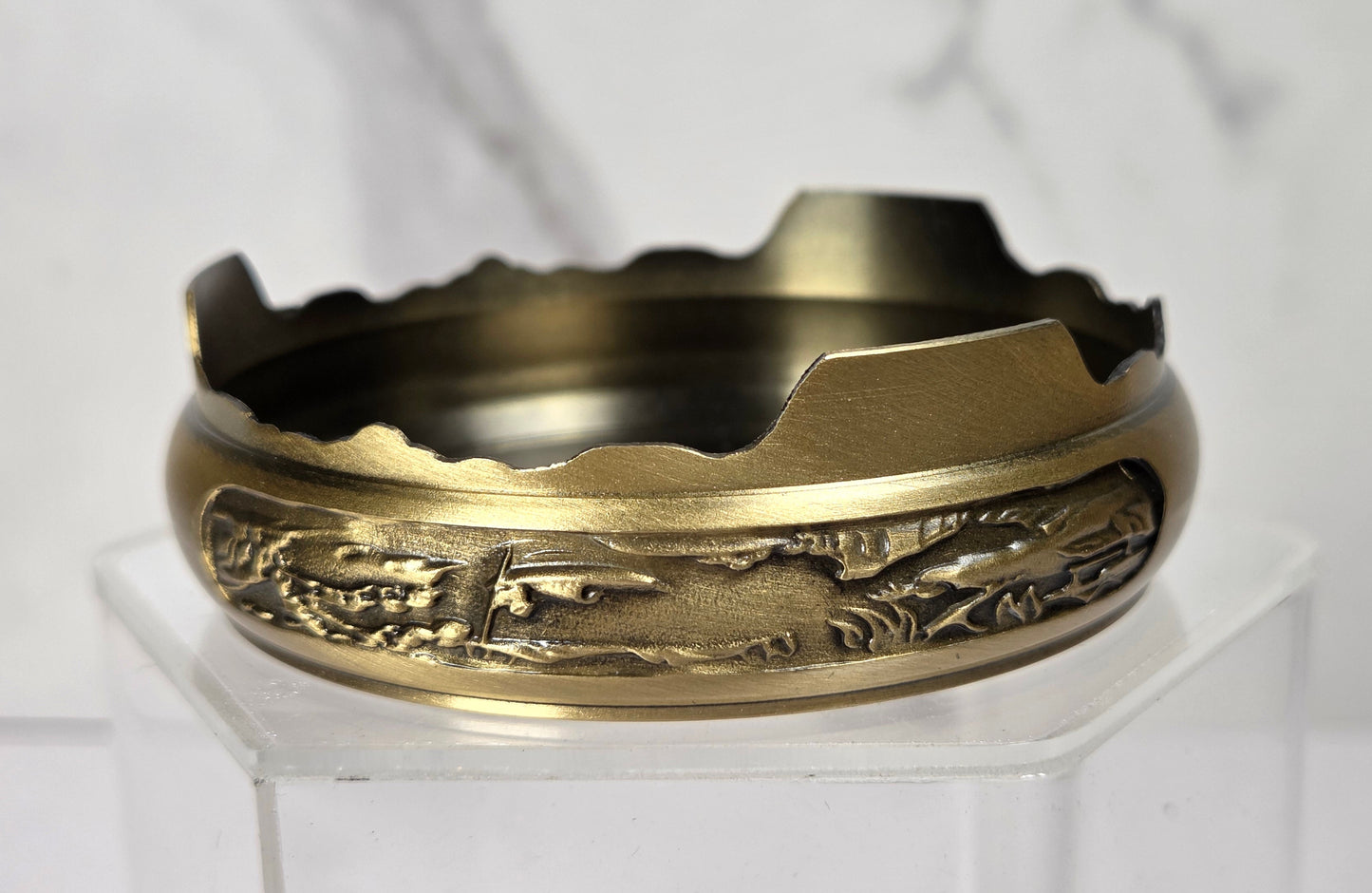 A vintage brass cigarette case lid with detailed embossed floral and scrollwork patterns, displayed on a clear acrylic stand against a white marble background.