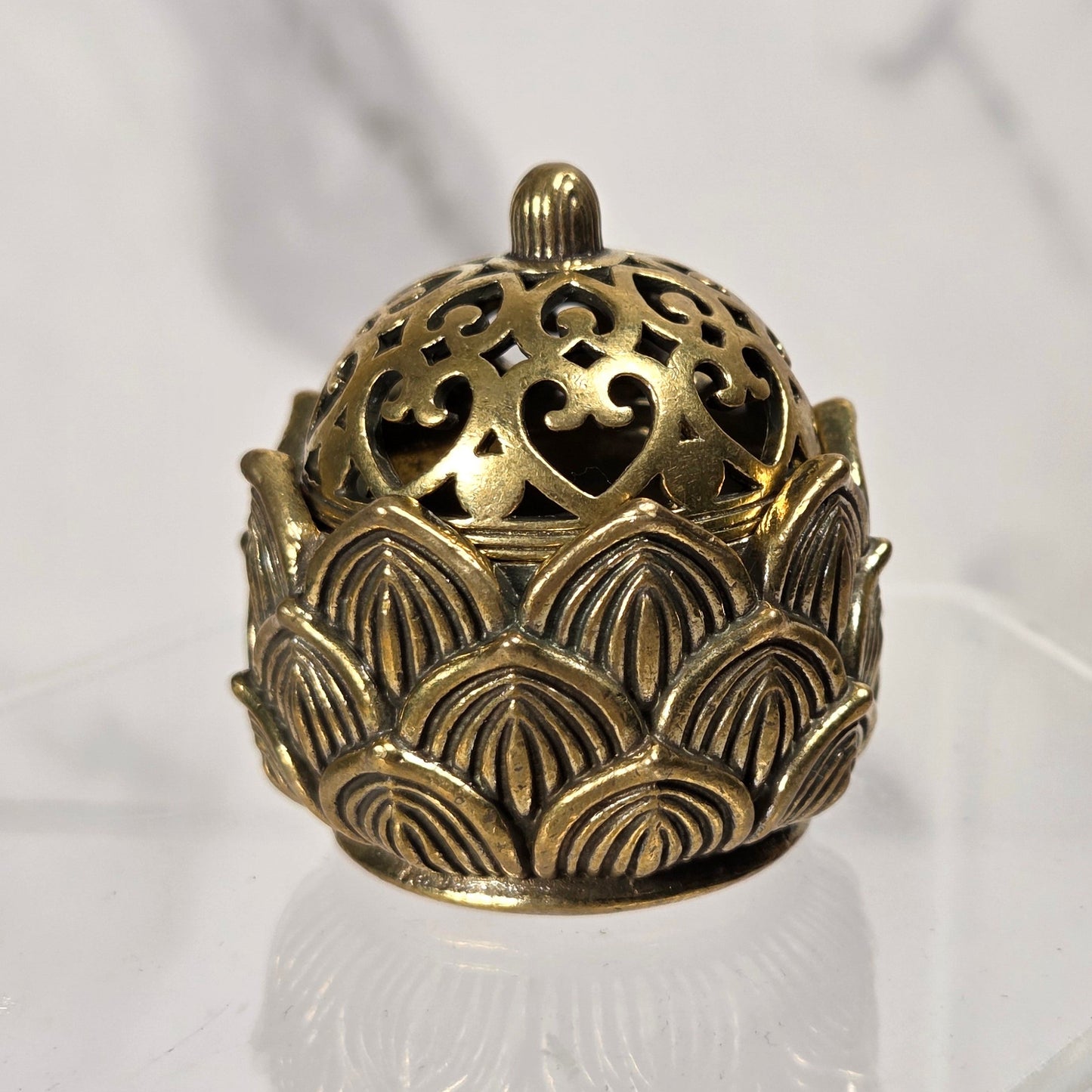 An ornate brass incense burner with a perforated lid and lotus-shaped base, displayed on a clear acrylic stand against a white marble background.