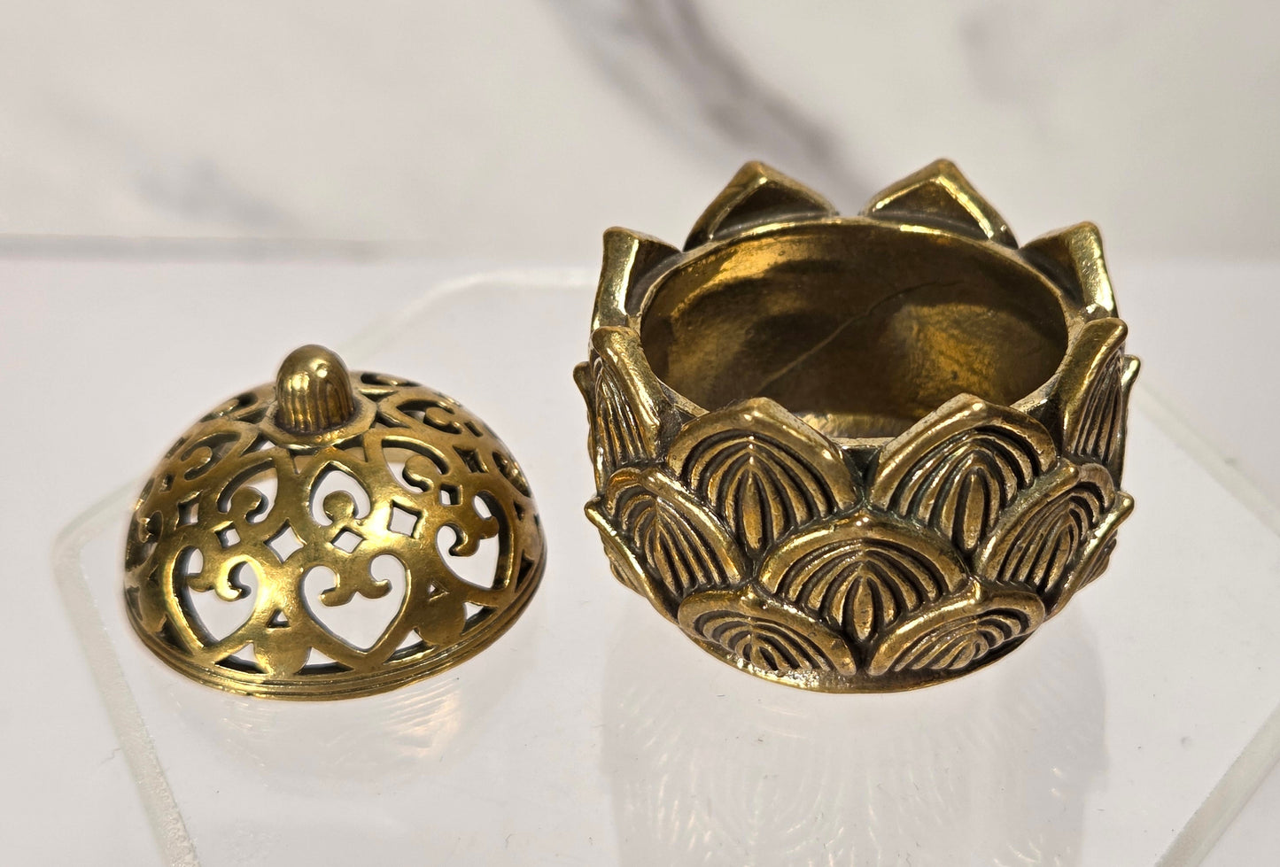 A small antique-style brass lotus-shaped incense holder with its lid removed, displayed on a clear acrylic stand against a light marble-patterned background.
