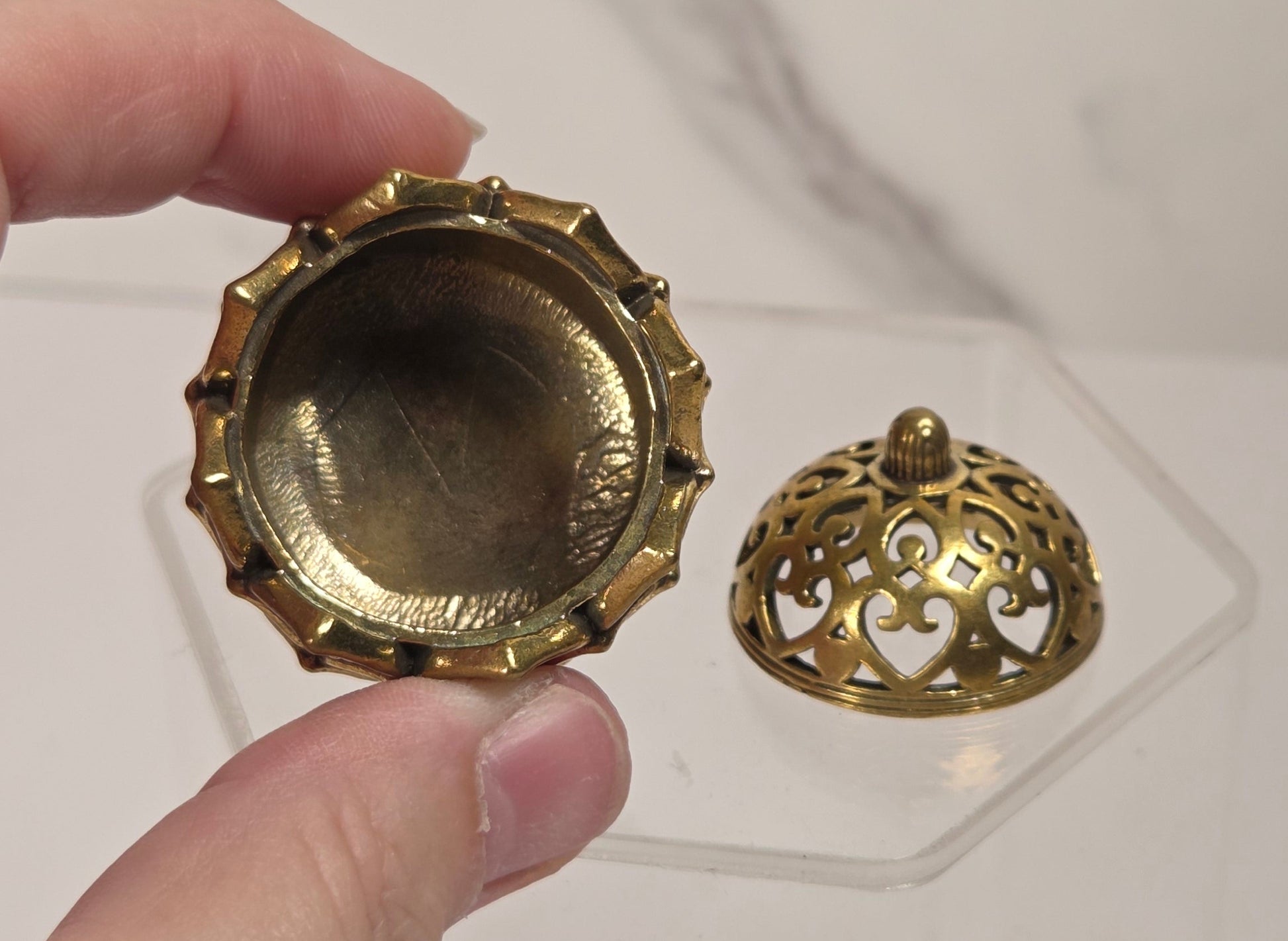A hand holds a small round brass-toned metal piece with a fluted edge next to a matching ornate dome-shaped lid on a clear acrylic stand, against a light marble-patterned background.