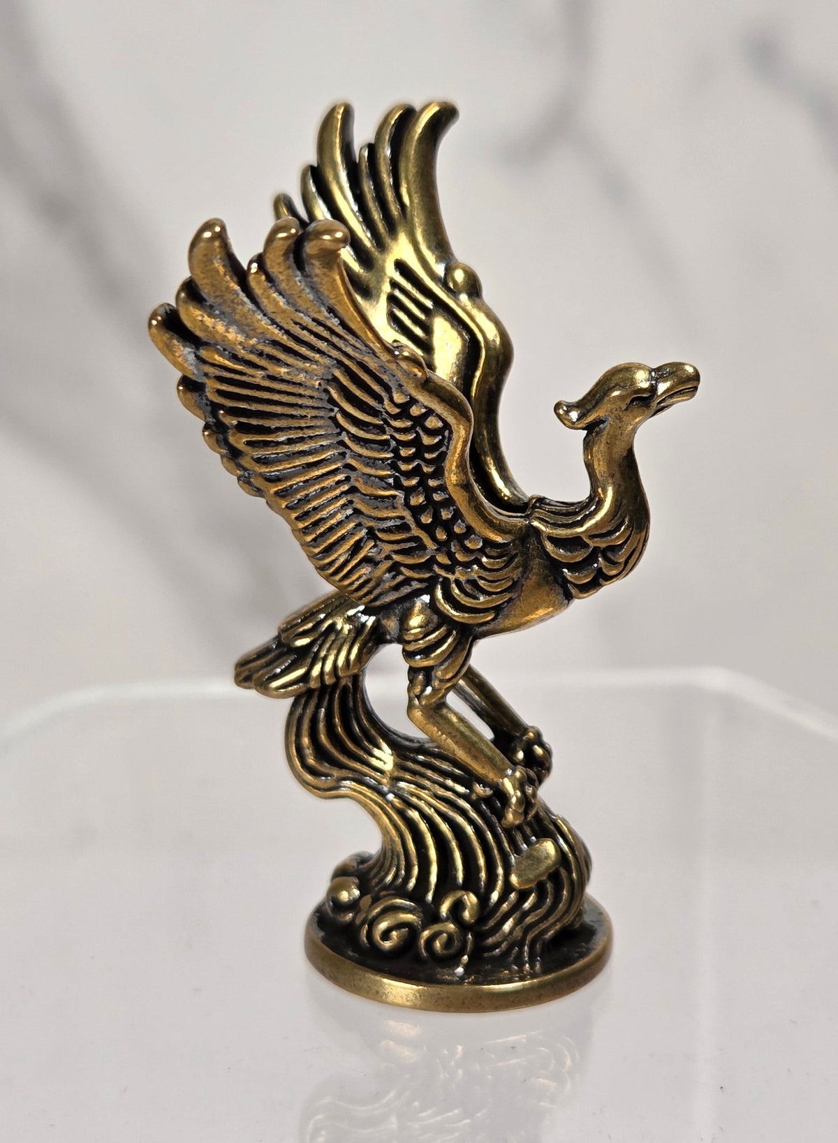 A small metallic decorative figurine of a phoenix or crane with wings raised, standing on a wave-like base. The object has an antique gold/brass finish and is displayed on a clear acrylic stand against a light marble-patterned background.