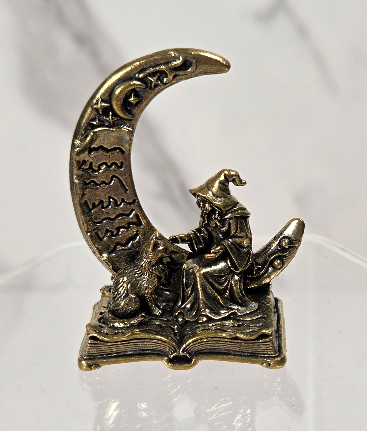 A small antique-style metal figurine of a wizard sitting on an open book and holding a wand, posed in front of a crescent moon, displayed on a clear acrylic stand.