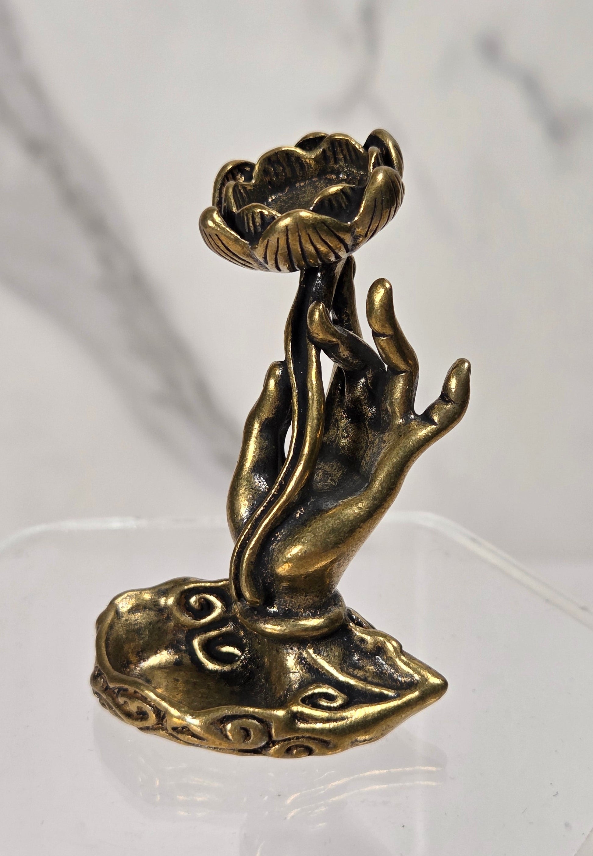 A small bronze-toned decorative figurine shaped like a lotus flower rising from a cloud-like base, displayed on a clear acrylic stand against a light marble-pattern background.