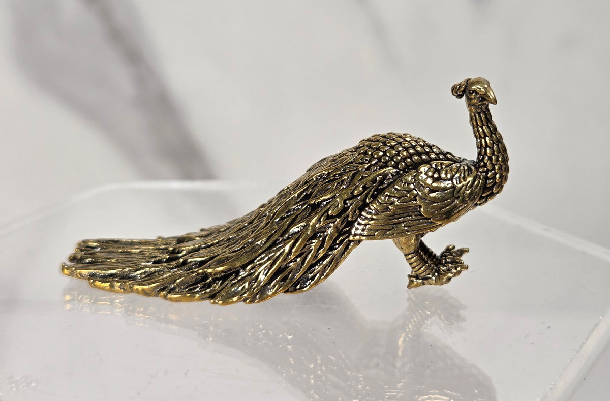 A small metallic peacock figurine in a textured gold finish, displayed on a clear acrylic stand against a light gray marble-patterned background.