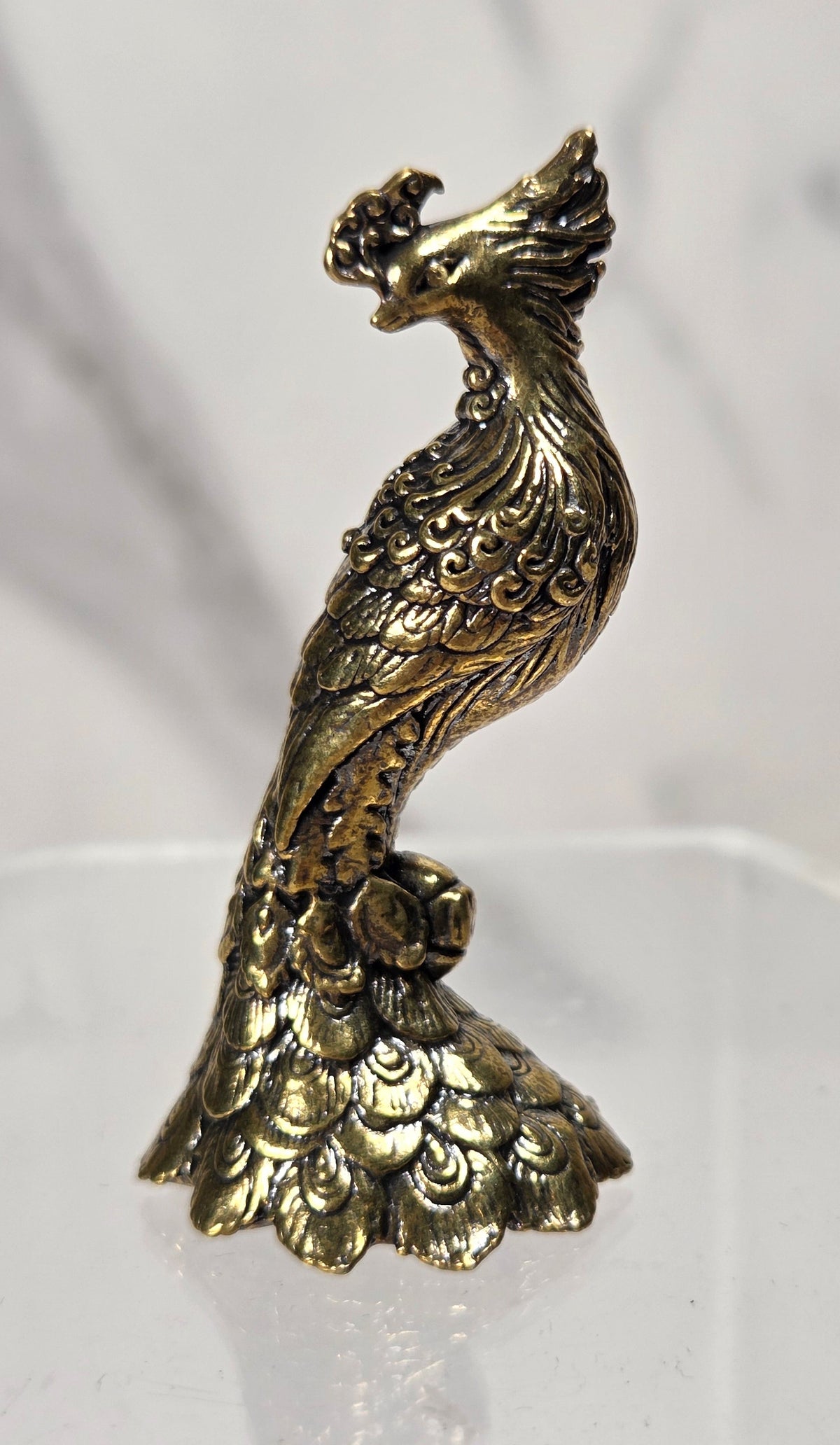 A small ornate metallic figurine shaped like a peacock, with an antique gold/brass finish, displayed on a clear acrylic stand against a light gray marble-patterned background.