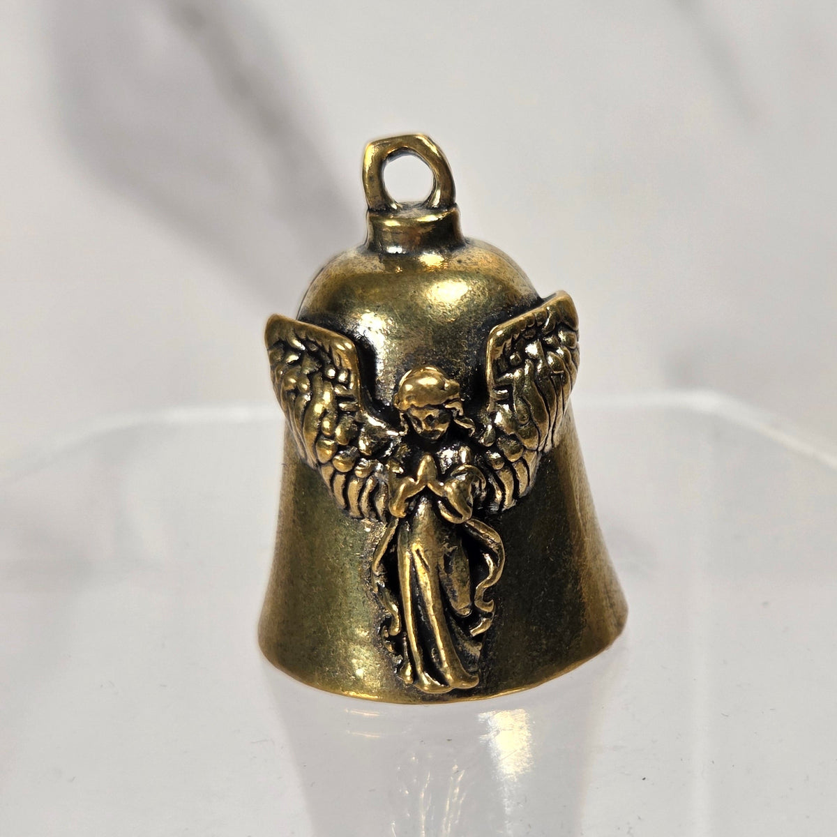 A small antique bronze angel bell charm with a detailed winged angel figure, displayed on a clear acrylic stand against a light gray marble-patterned background.