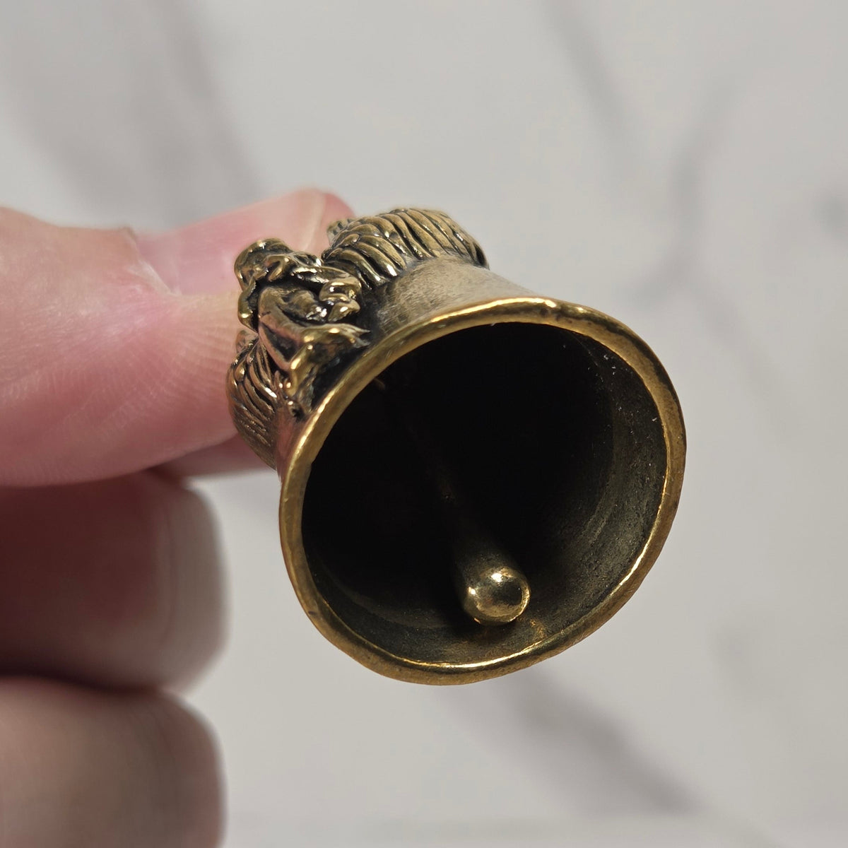 A hand holding a small antique-style brass bell with a lion head finial against a light gray marble-tiled background.