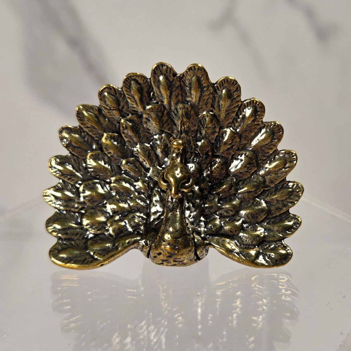 A small metallic gold peacock figurine with a textured finish, displayed on a clear acrylic stand against a light gray marble-patterned background.